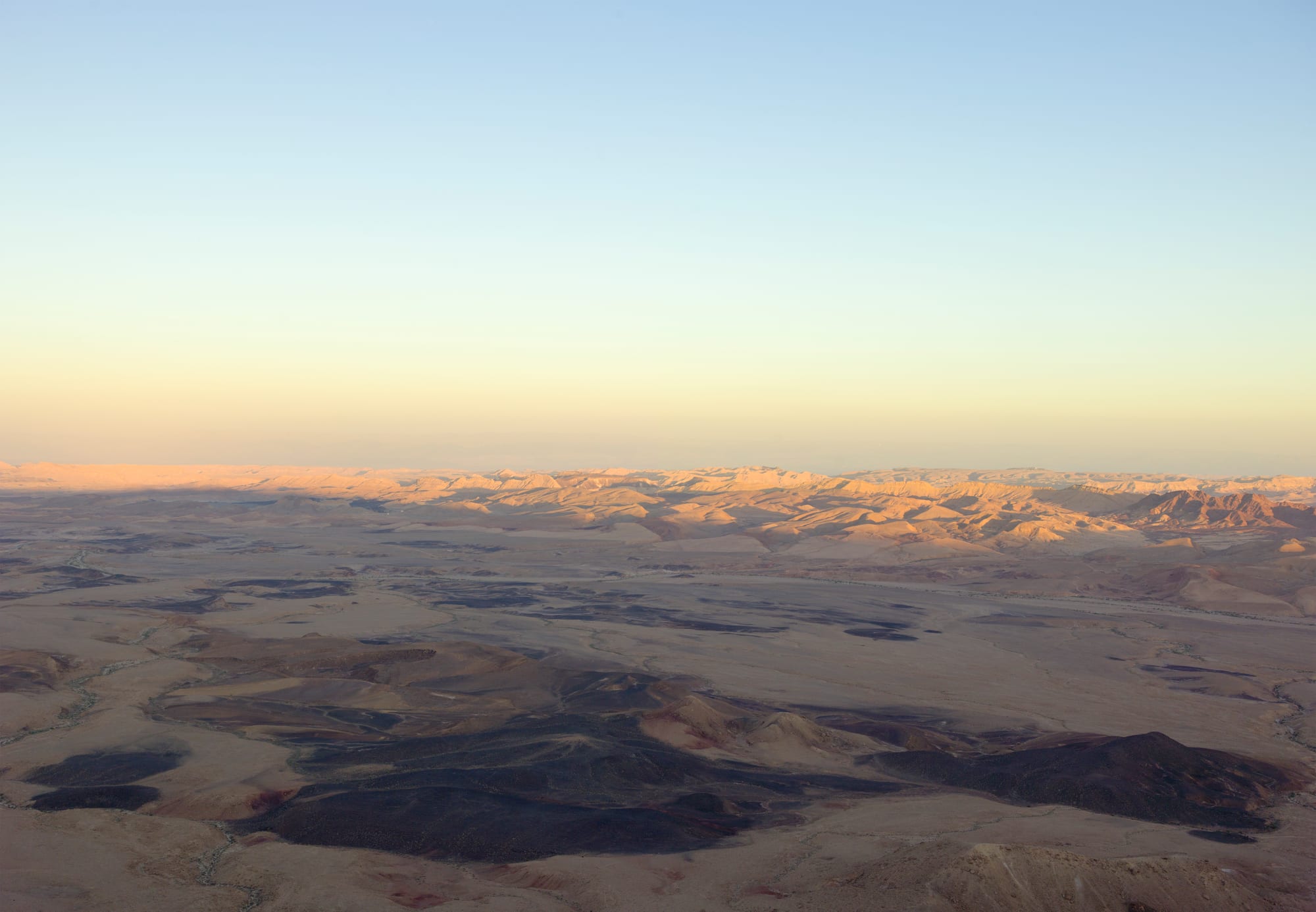 A vast desert basin of pale sand and dark rock outcrops, with rows of low ridges glowing in soft evening light