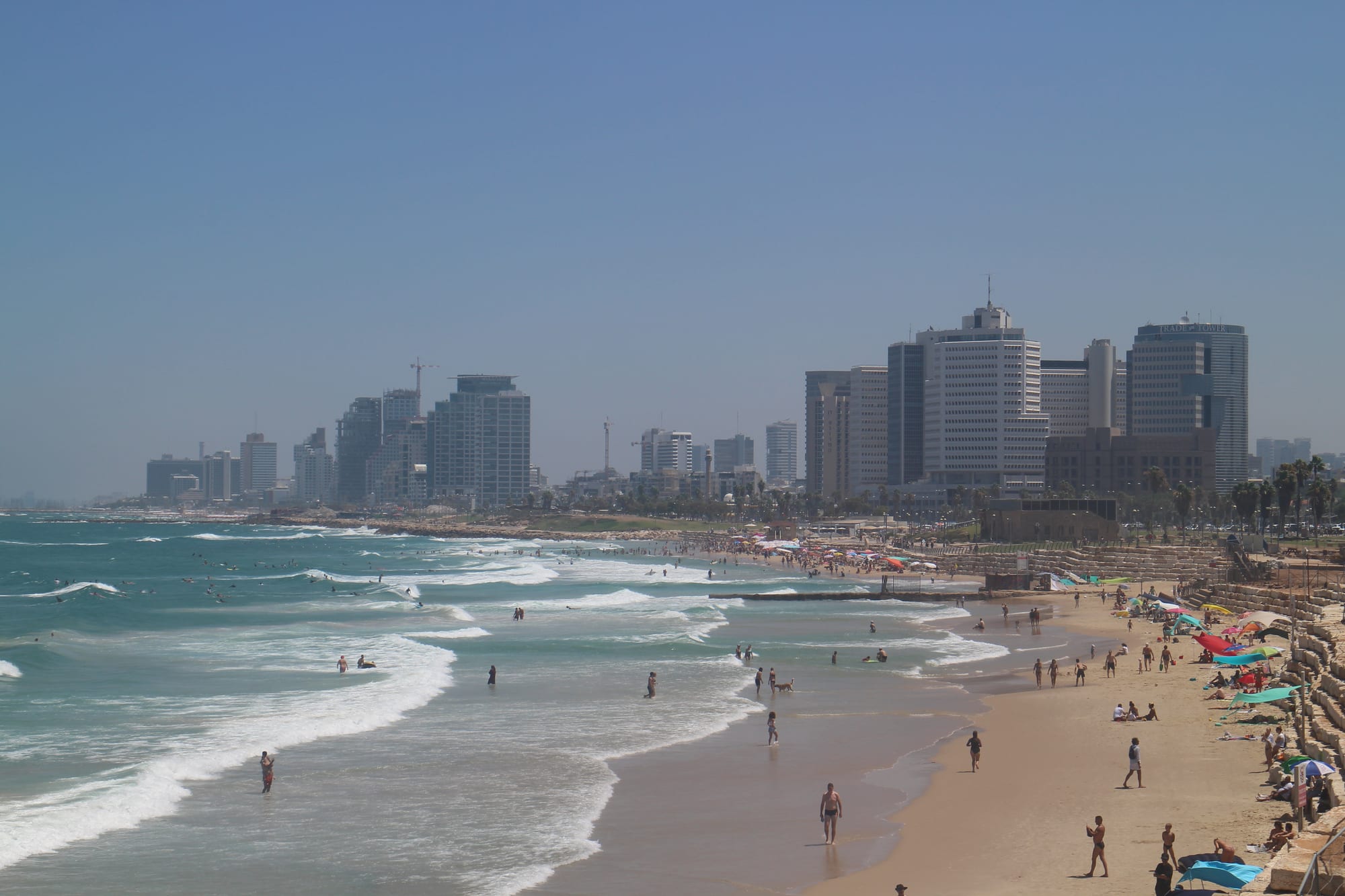 A cluster of glass office and residential towers rising above lower white buildings along a Mediterranean coastline