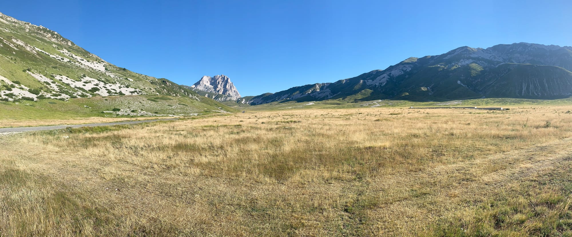 A wide golden grassland plateau under a clear blue sky, with the rocky peak of Corno Grande and green ridges beyond