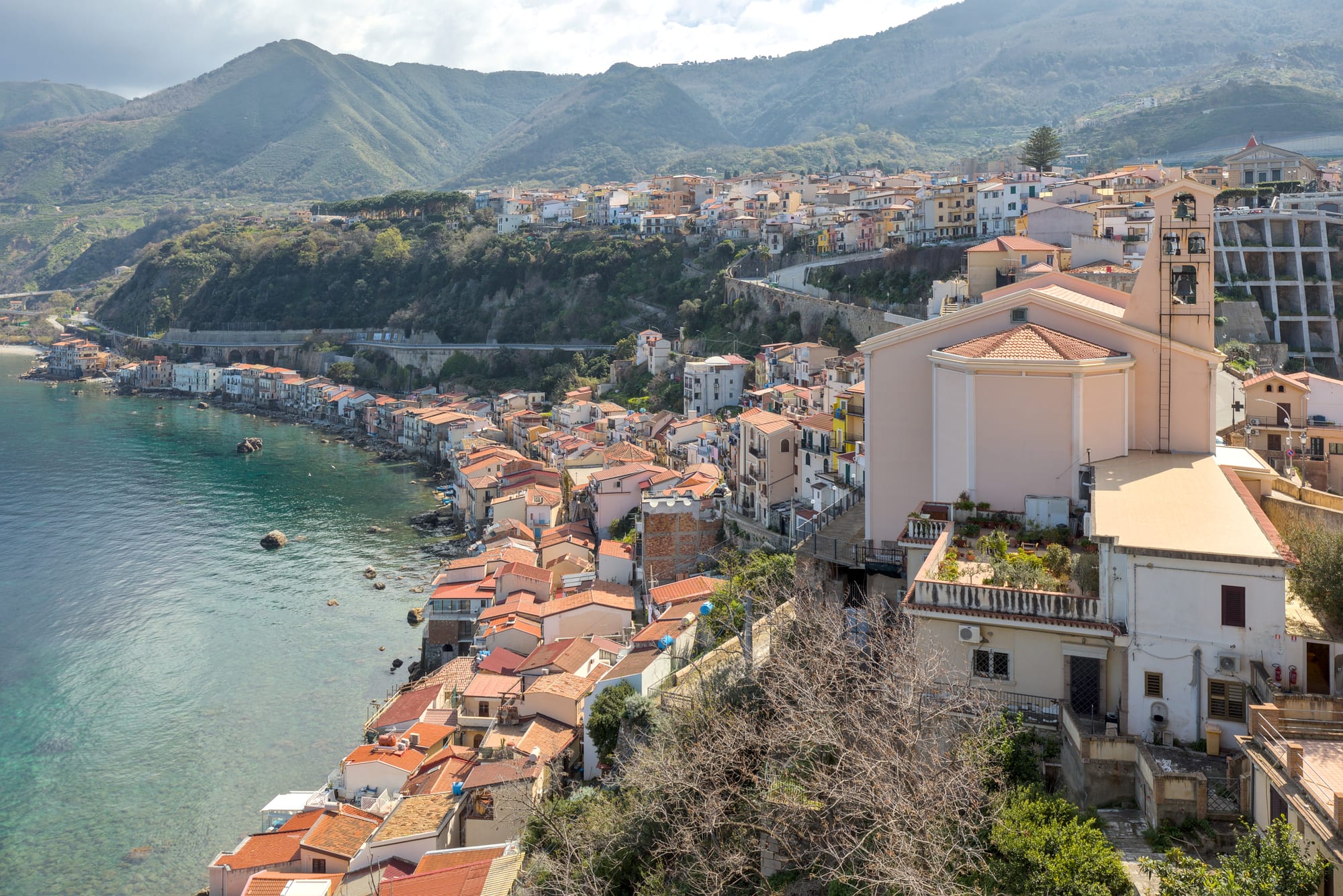 Tiled houses of a fishing quarter lining clear green water, with a church and wooded mountains behind