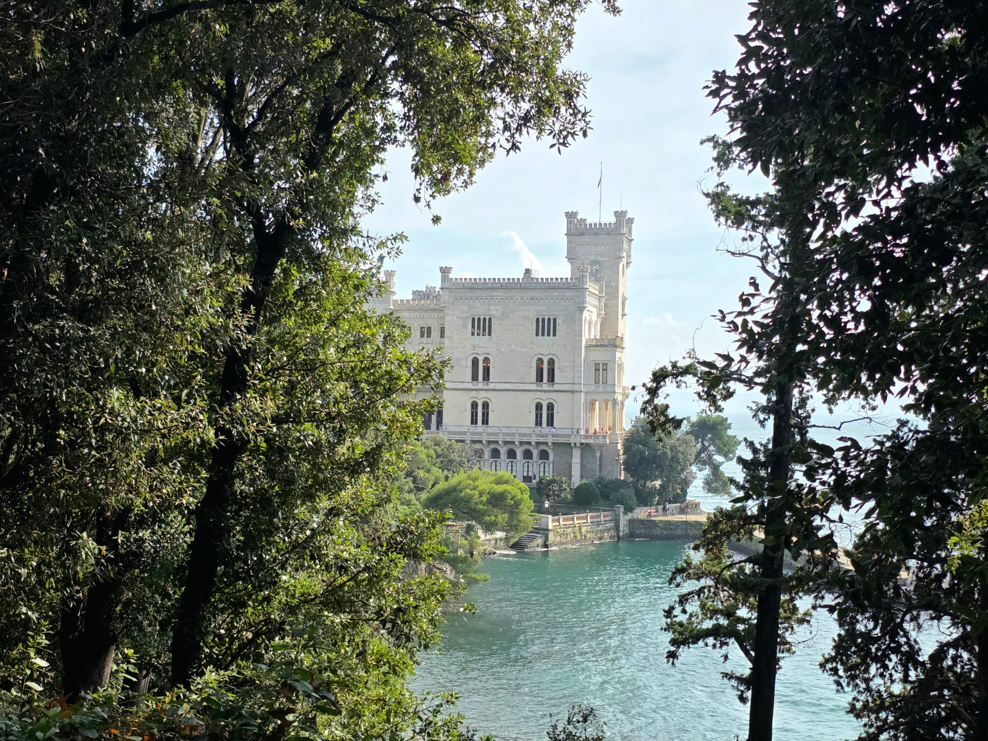 A white castellated castle on a rocky point above turquoise water, framed by dark trees