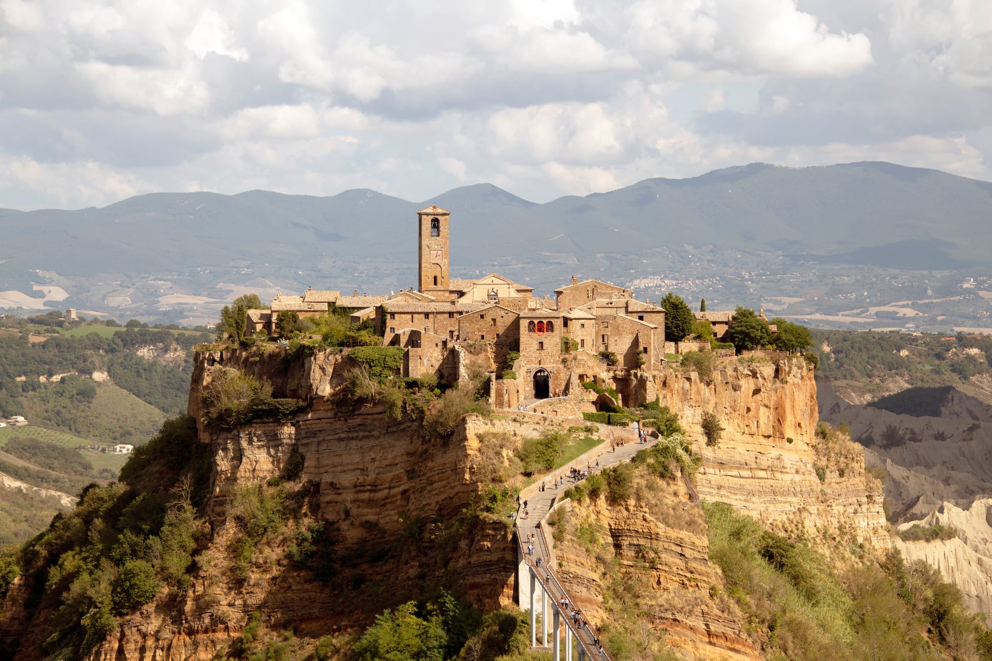 The stone village of Civita di Bagnoregio on an eroded cliff-top, reached by a long footbridge, with hills behind