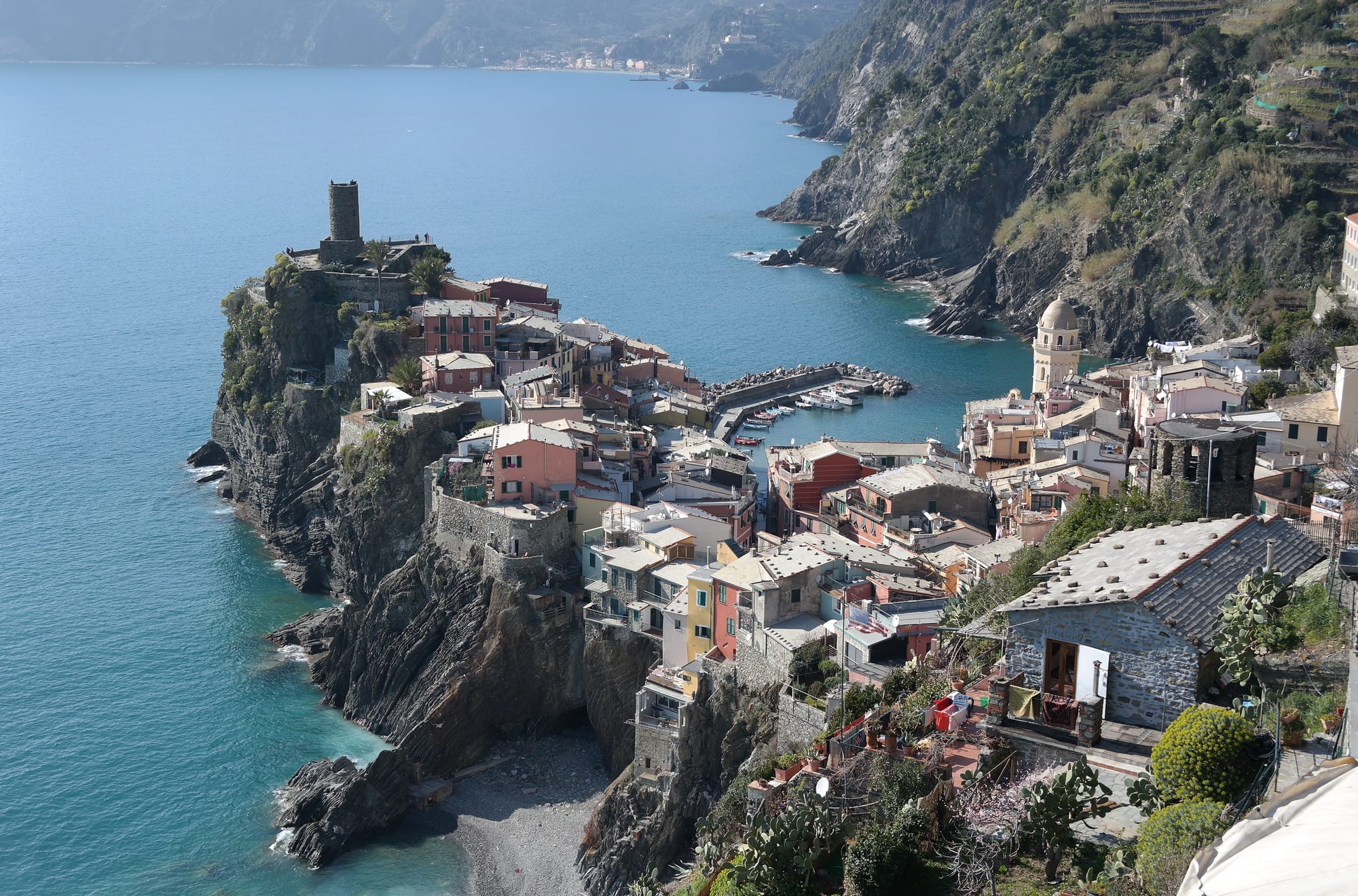 The coloured houses and small harbour of Vernazza on a rocky point, with a castle tower and steep terraced cliffs above the sea