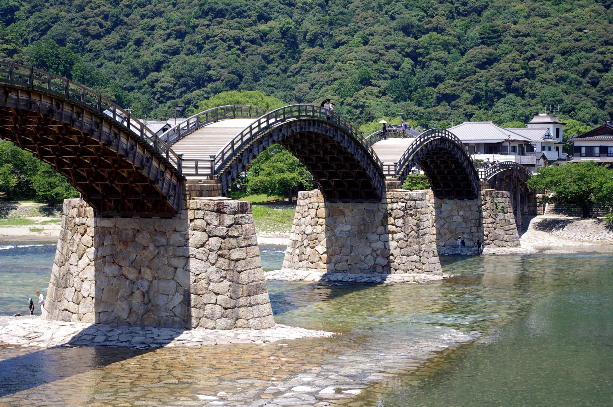 A wooden bridge of steep arches resting on stone piers across a clear, shallow river below wooded hills