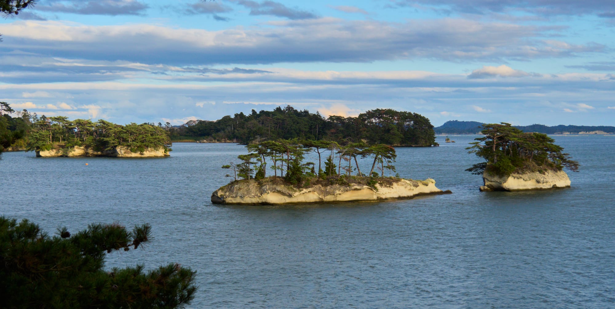 Small rocky islets topped with pine trees scattered across a calm blue bay under a cloudy evening sky