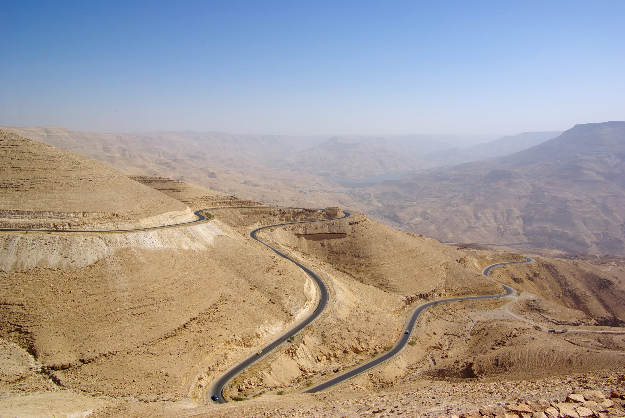 A road winding in hairpin bends down the bare, pale slopes of a wide desert canyon, with a reservoir in the haze below