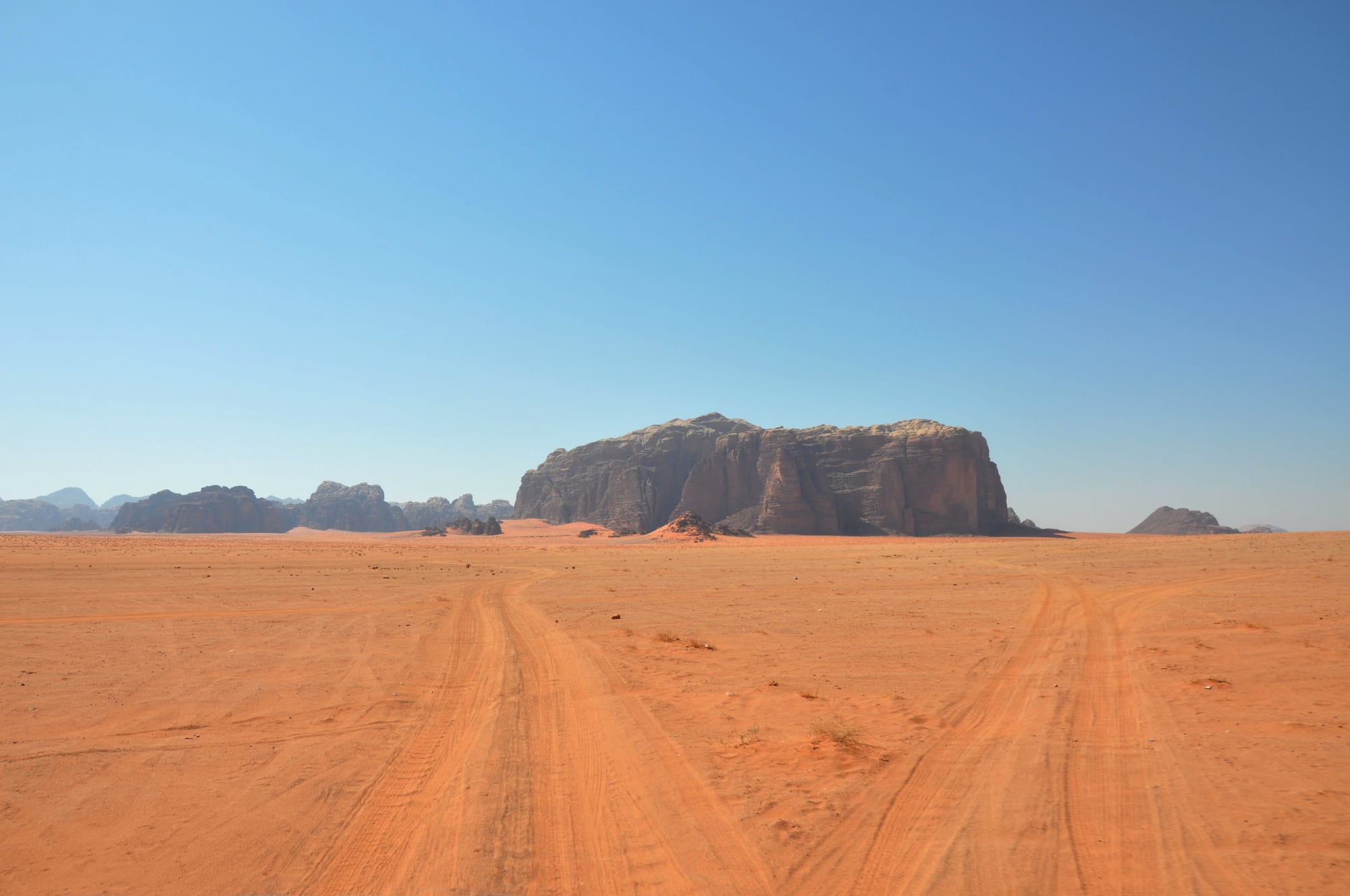 Tyre tracks crossing flat orange sand towards a massive sandstone outcrop under a clear blue sky