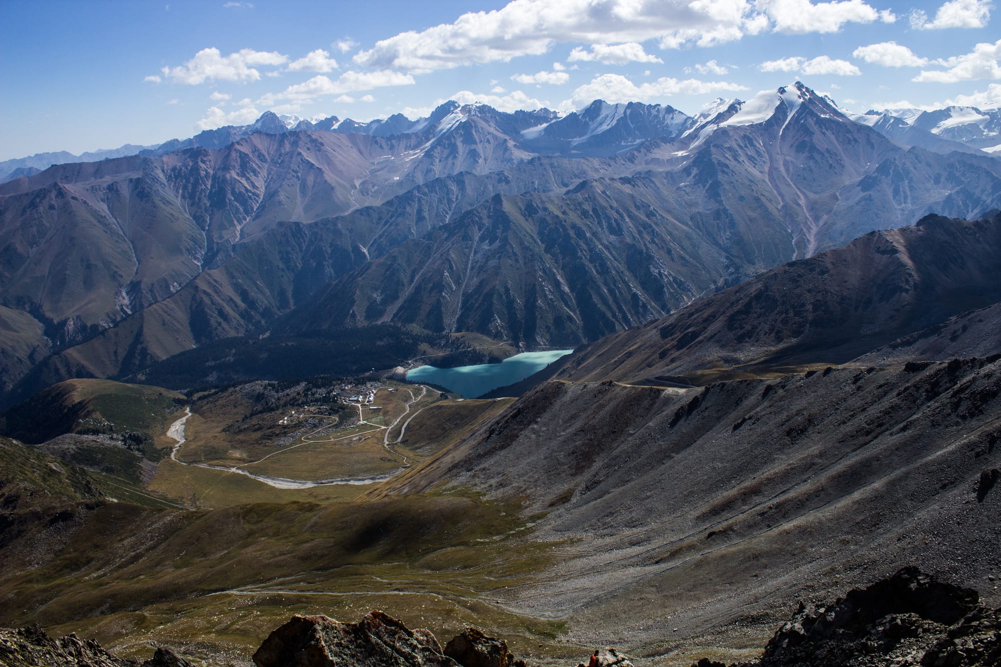 A turquoise lake in a green valley far below bare grey mountain slopes, with snow-capped peaks along the skyline