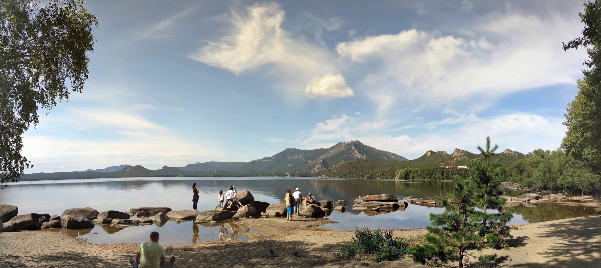 A calm lake with boulders along a sandy shore, pine-covered hills and a rocky peak rising beyond under a blue sky