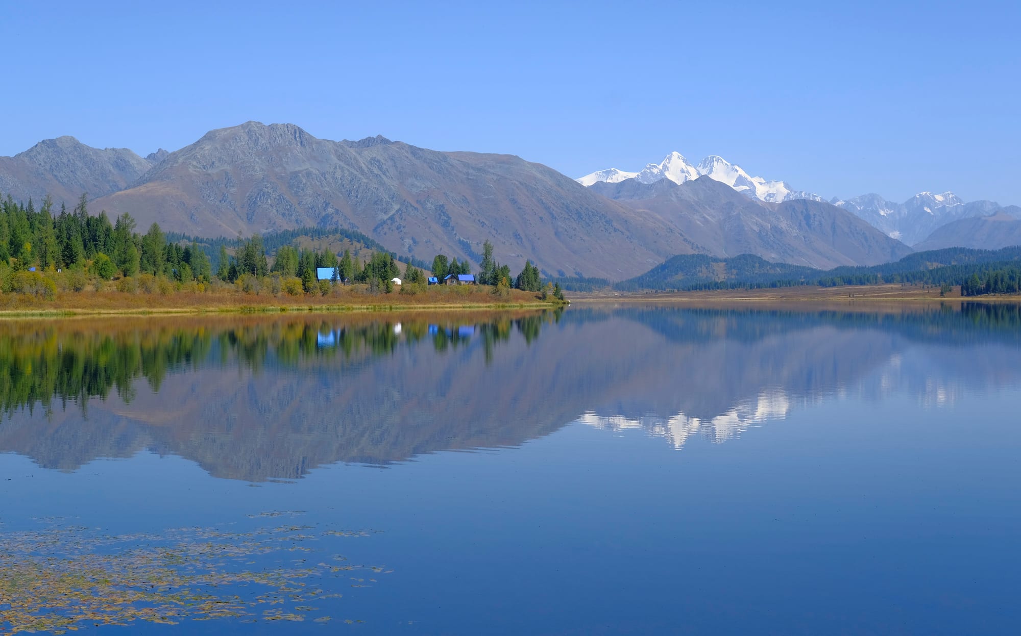A still lake reflecting forested shores, grey mountain slopes and a snow-covered twin peak under a clear blue sky