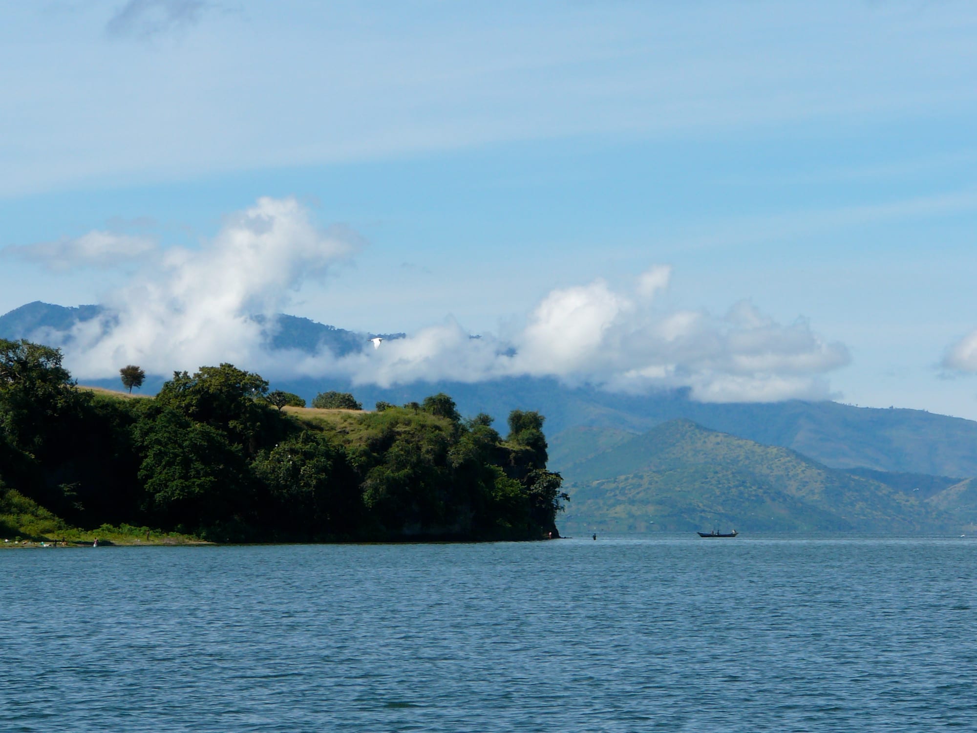 A wooded green headland on calm blue lake water, with cloud-capped hills behind and a small boat offshore