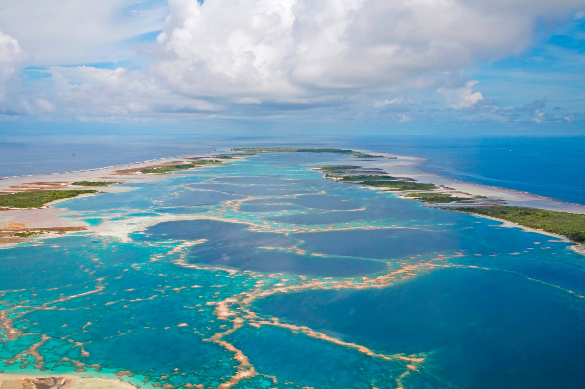 Aerial view of a long coral atoll, its deep-blue lagoon laced with pale orange reef ridges and turquoise shallows, ringed by low green islets under towering white clouds