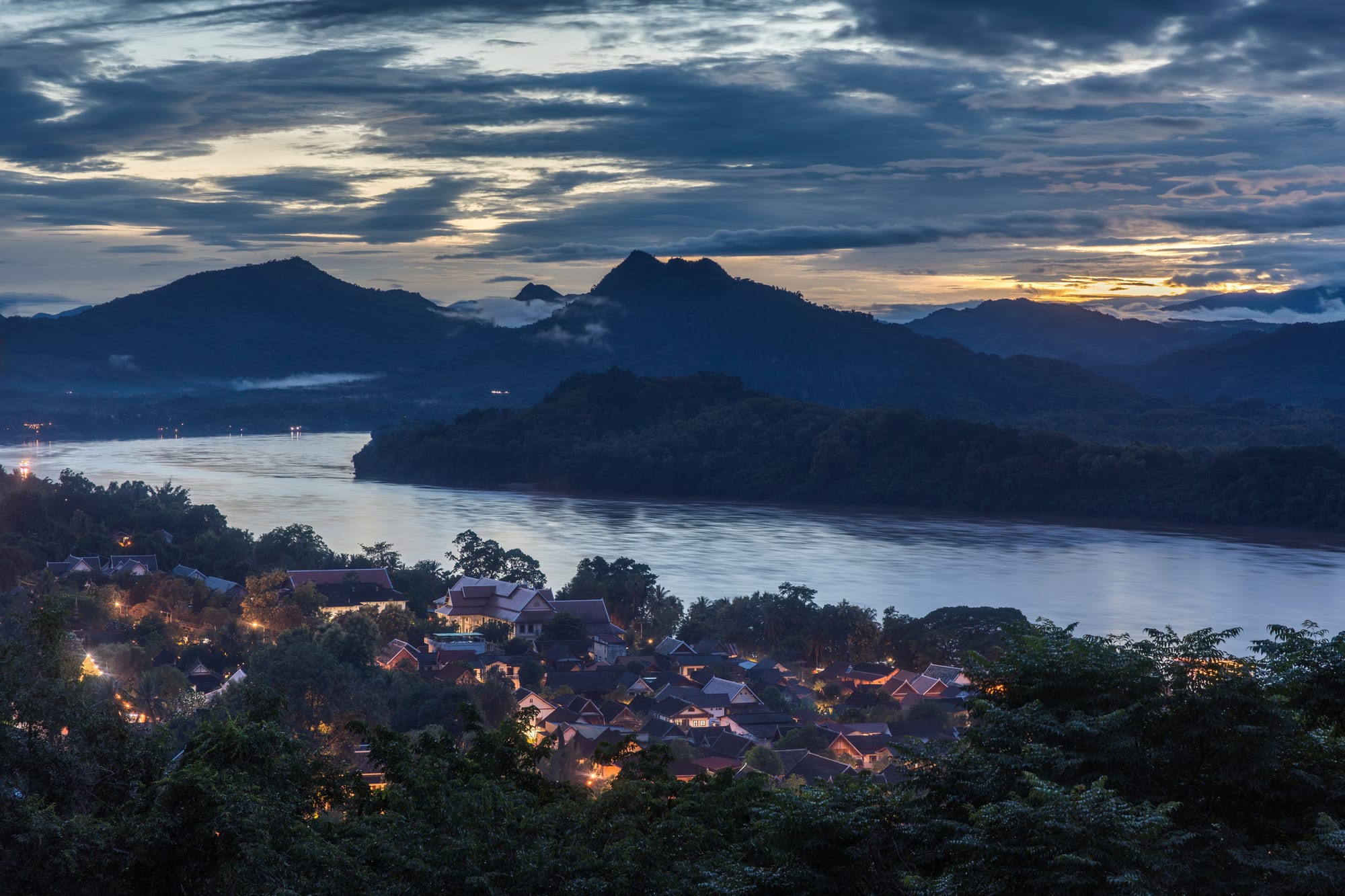 Dusk over the wide Mekong with forested hills and lit rooftops of Luang Prabang among trees in the foreground