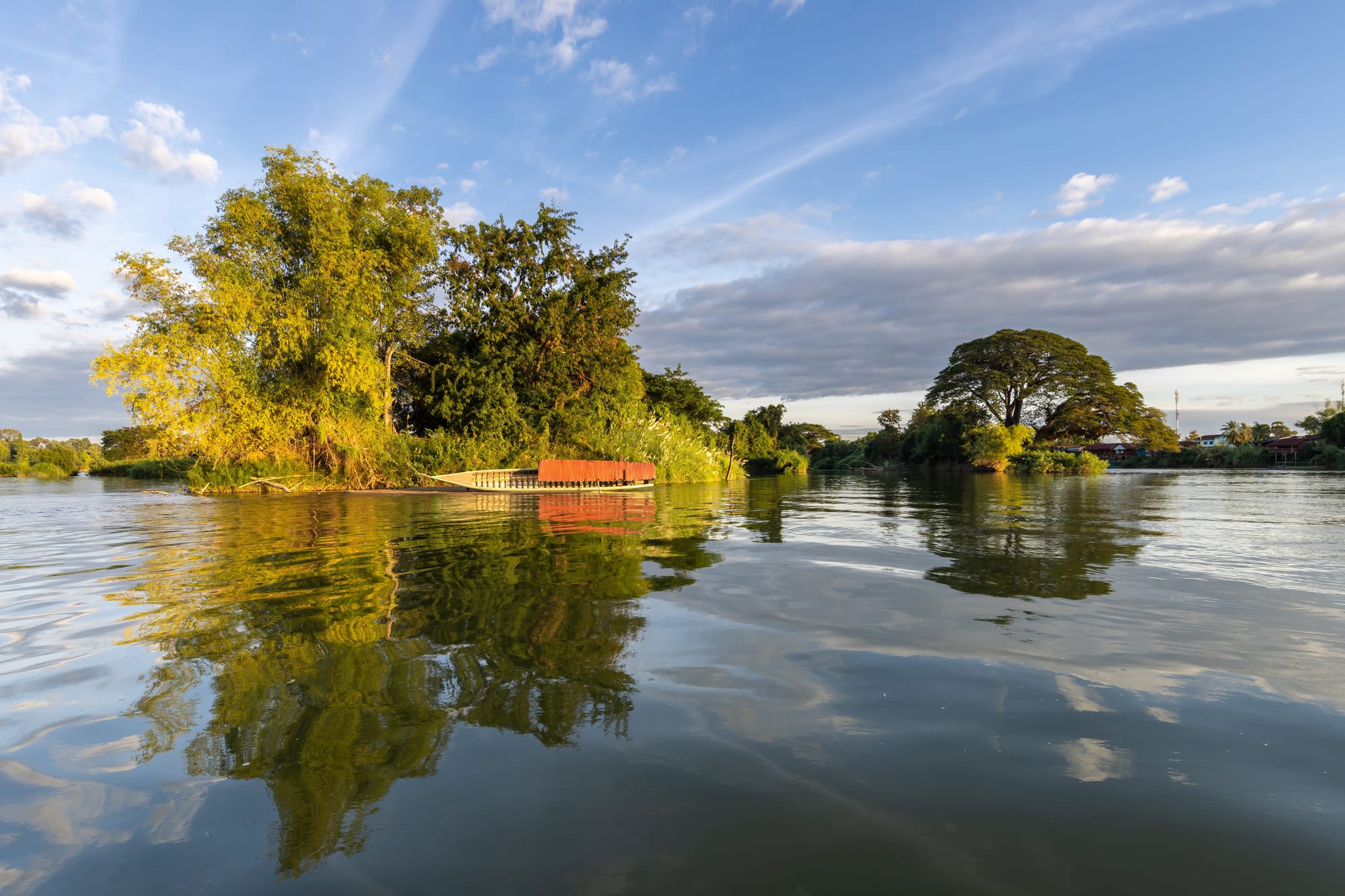 Wooded islands and a beached wooden boat reflected in the calm Mekong under a blue sky with evening light
