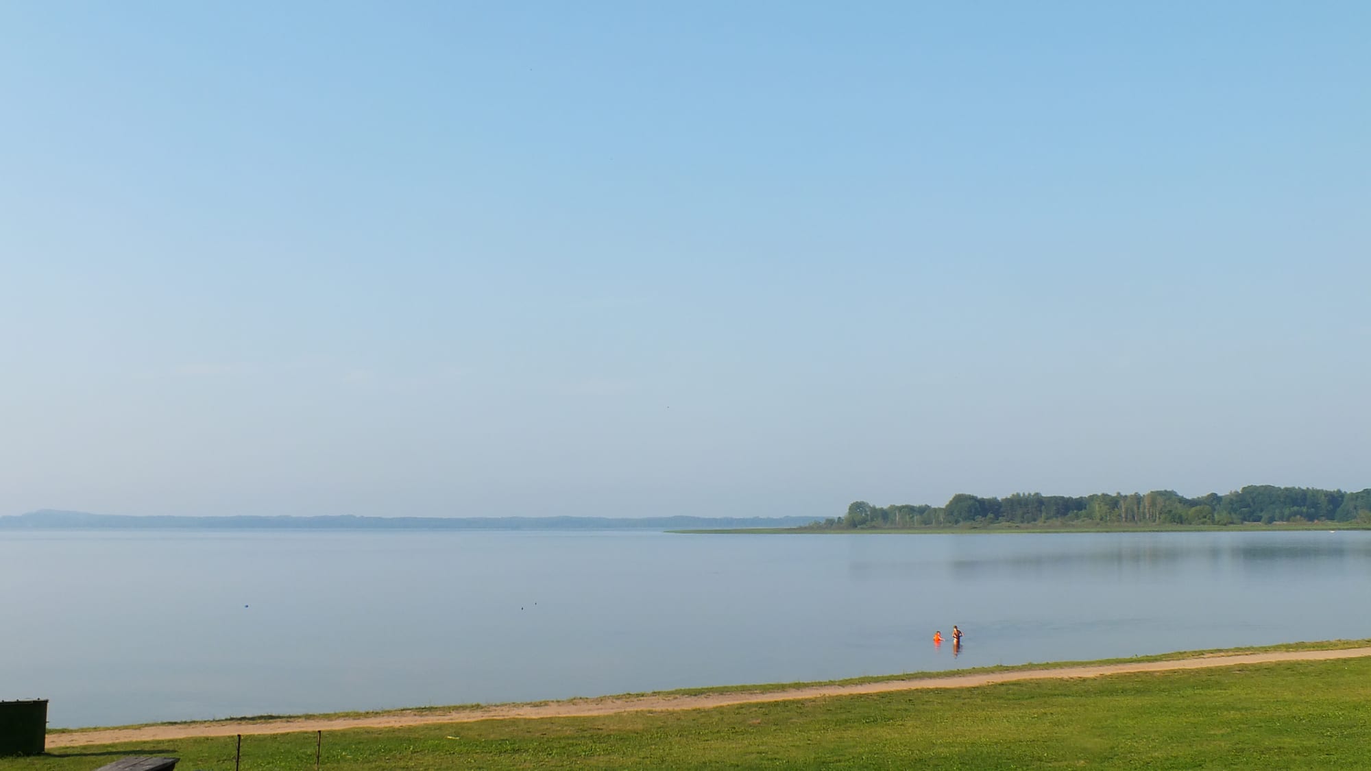 A wide, calm lake under a pale blue sky, with a wooded point on the right and a grassy shore in front