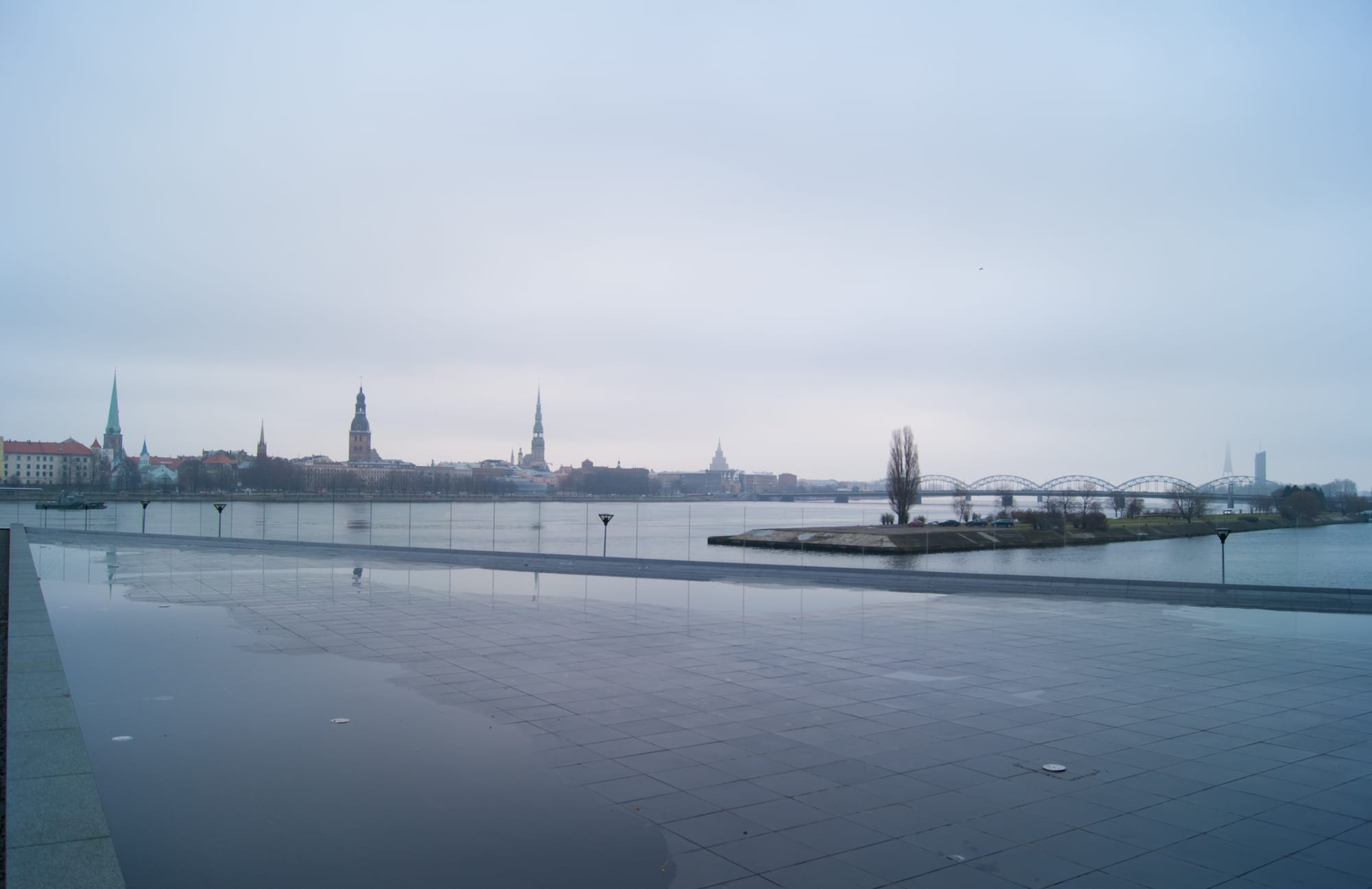 A skyline of church spires and red-tiled roofs rising above a river, seen from across the water under a pale sky