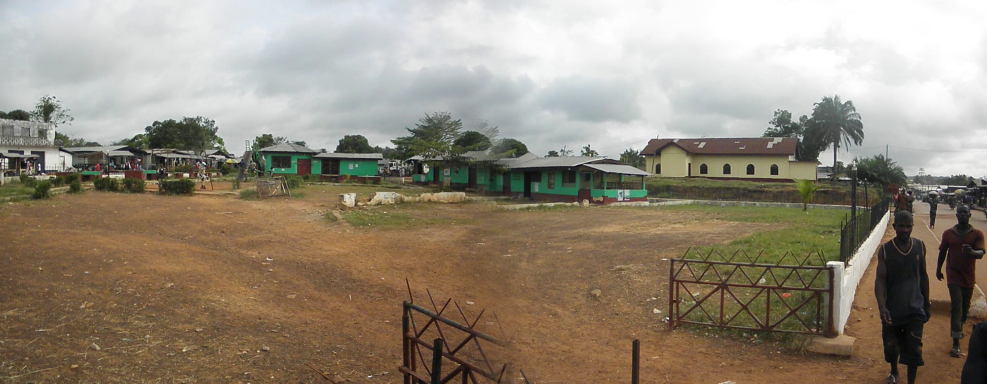 Single-storey green and white buildings around an open, reddish-earth yard, with palm trees under a cloudy sky