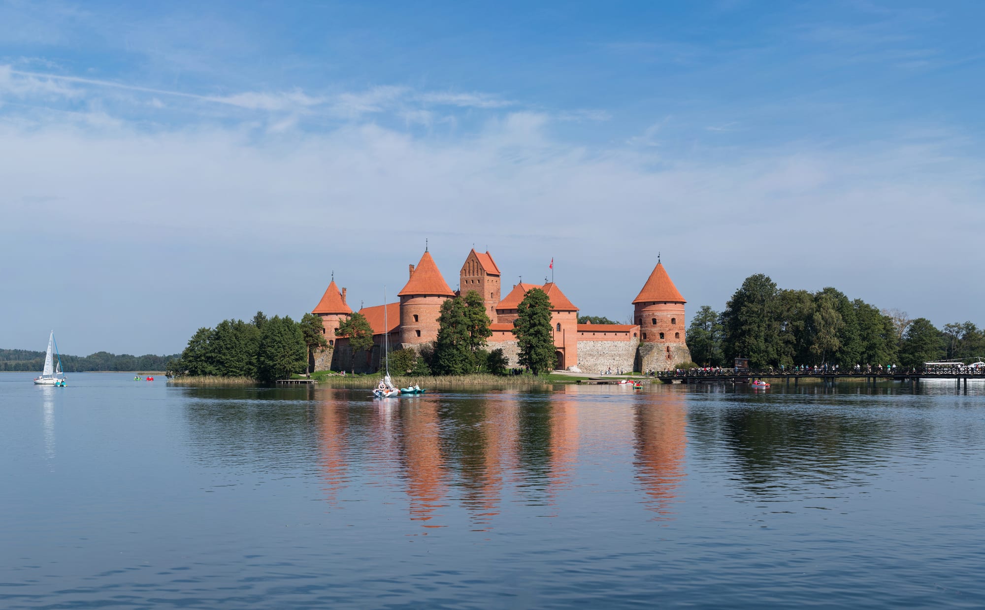A red-brick castle with towers and a keep on a small island in a calm lake, connected to the shore by a footbridge