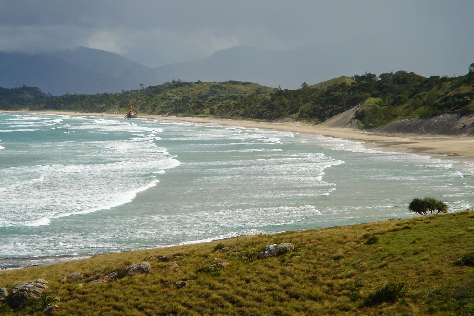 Surf rolling onto a long sandy beach below grassy slopes, with a shipwreck offshore and cloud-covered mountains behind