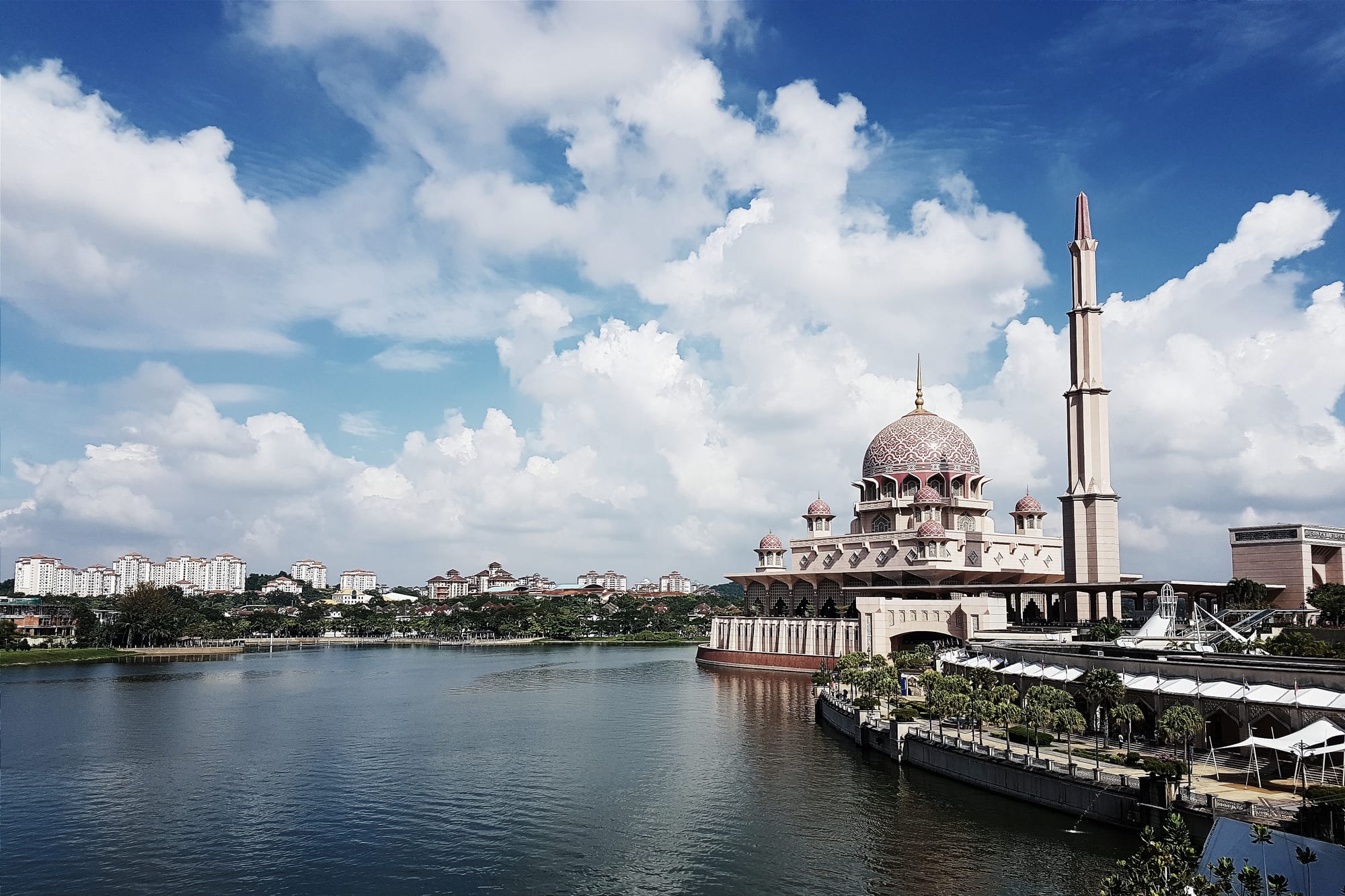 A domed pink mosque with a tall minaret on the edge of a wide lake under white clouds, apartment blocks on the far shore