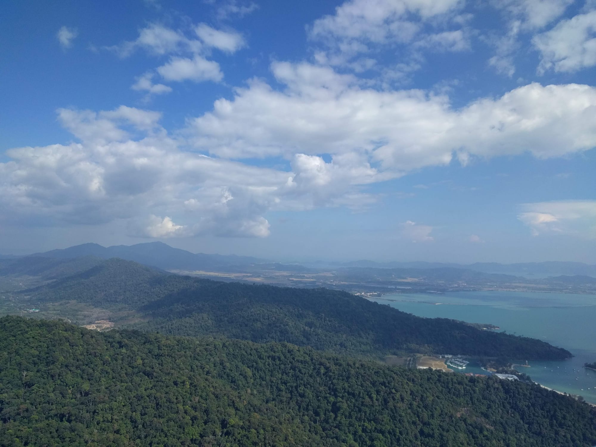 Forested hills falling to a turquoise bay under a blue sky with white clouds, seen from a high summit