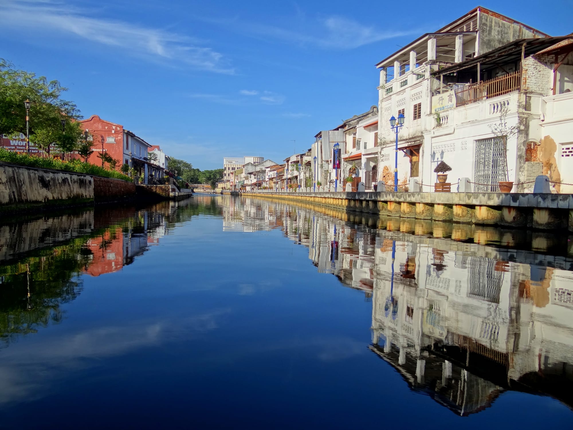 A calm river reflecting old white and red shophouses and a blue sky, with a riverside walkway along the bank