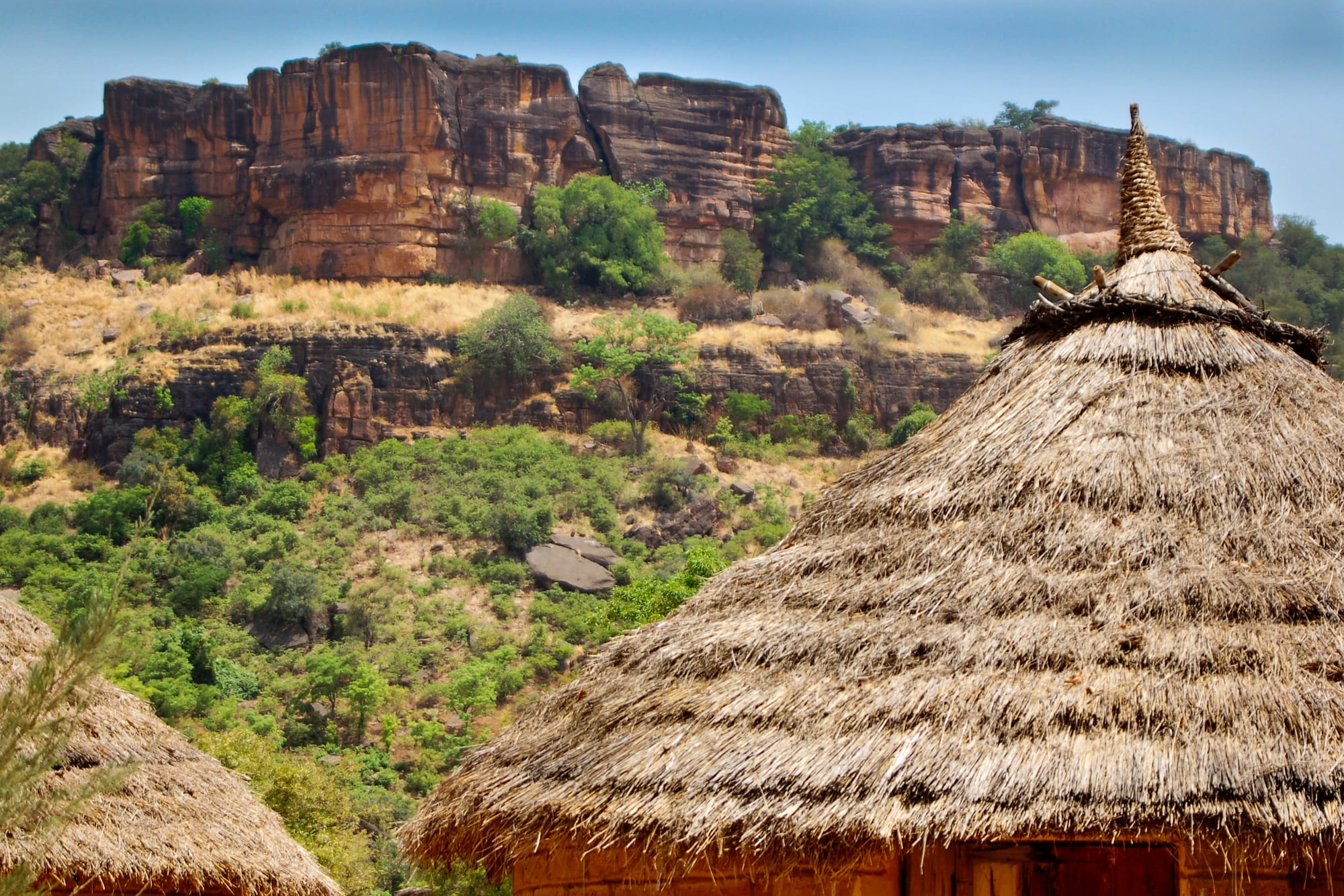 Red sandstone cliffs above a wooded green slope, with the thatched conical roof of a round hut in the foreground