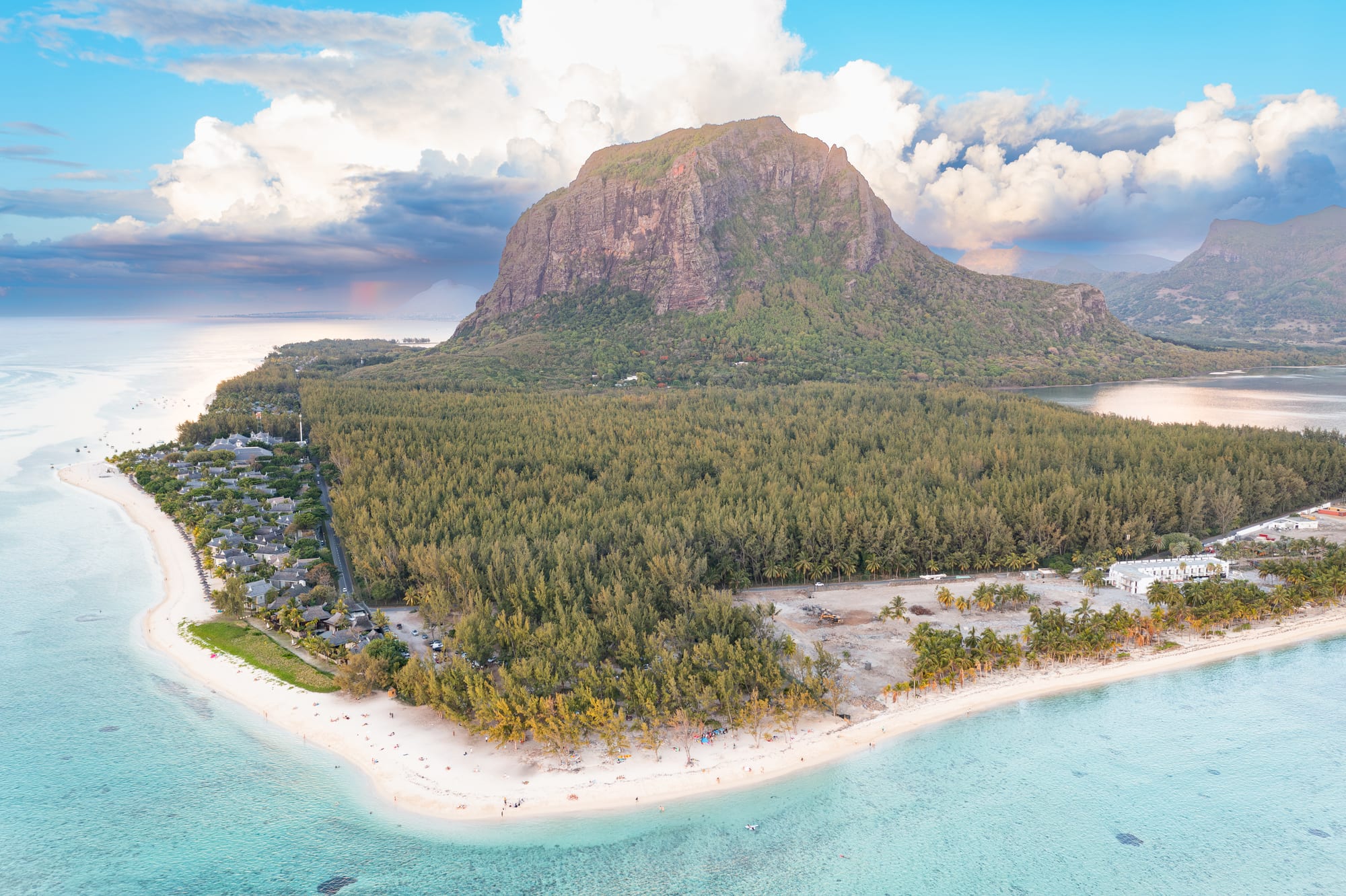 An aerial view of a dark basalt mountain on a green peninsula ringed by brilliant turquoise lagoon and pale reef