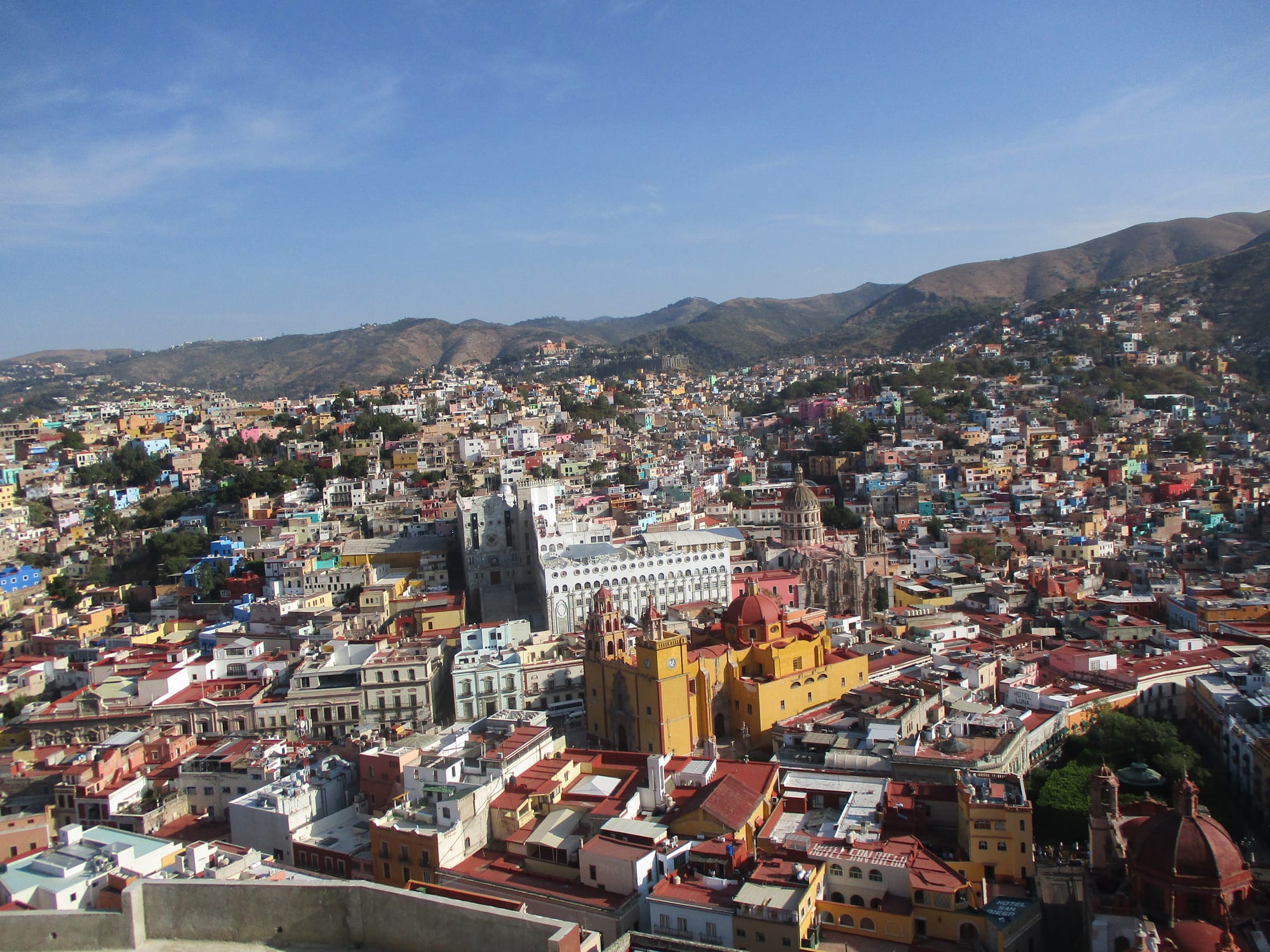 Brightly painted houses filling a valley below brown hills, with a yellow domed basilica and a white university building