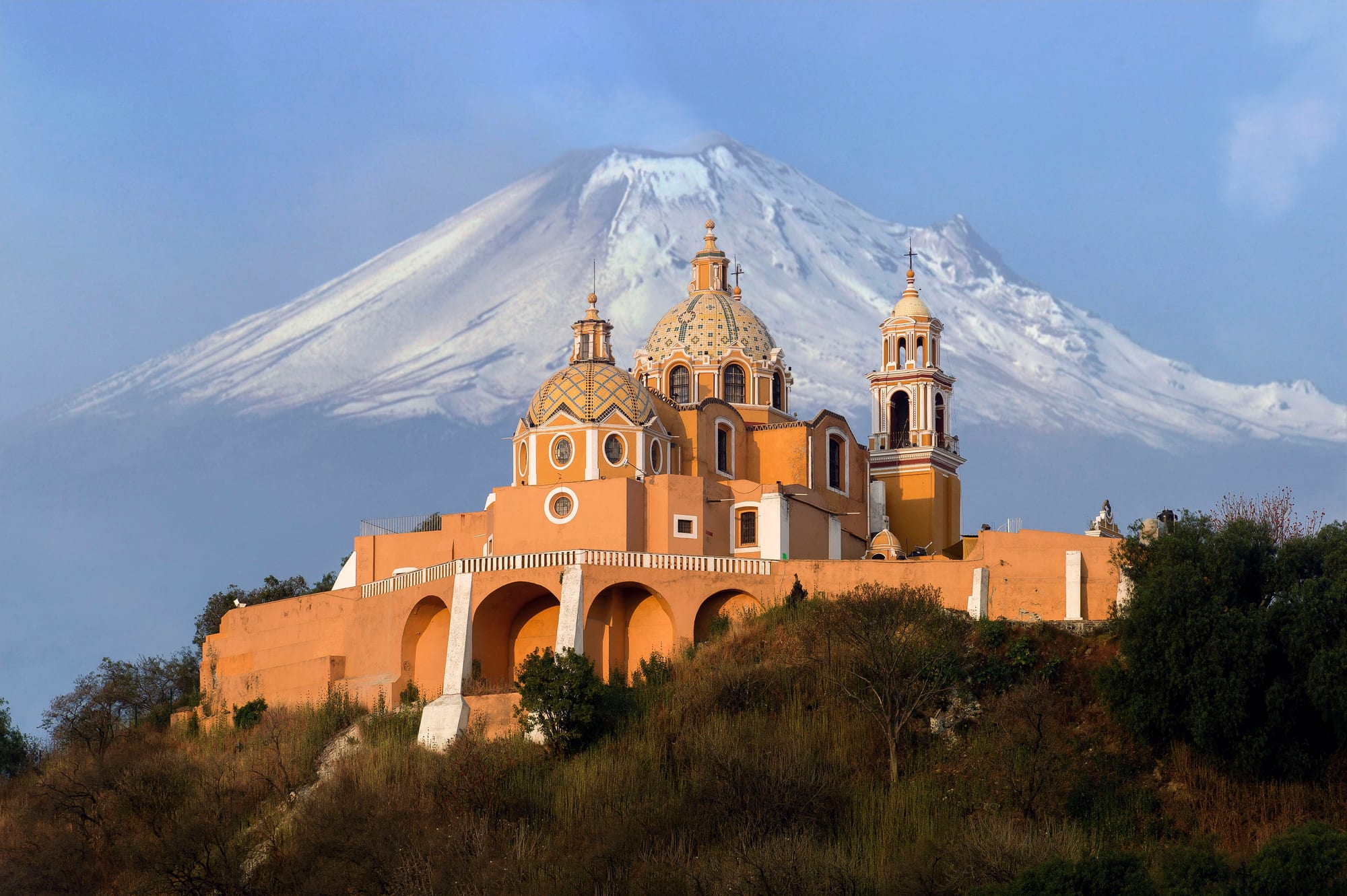 A yellow church with tiled domes on a wooded hilltop in front of a snow-covered volcano under a blue sky