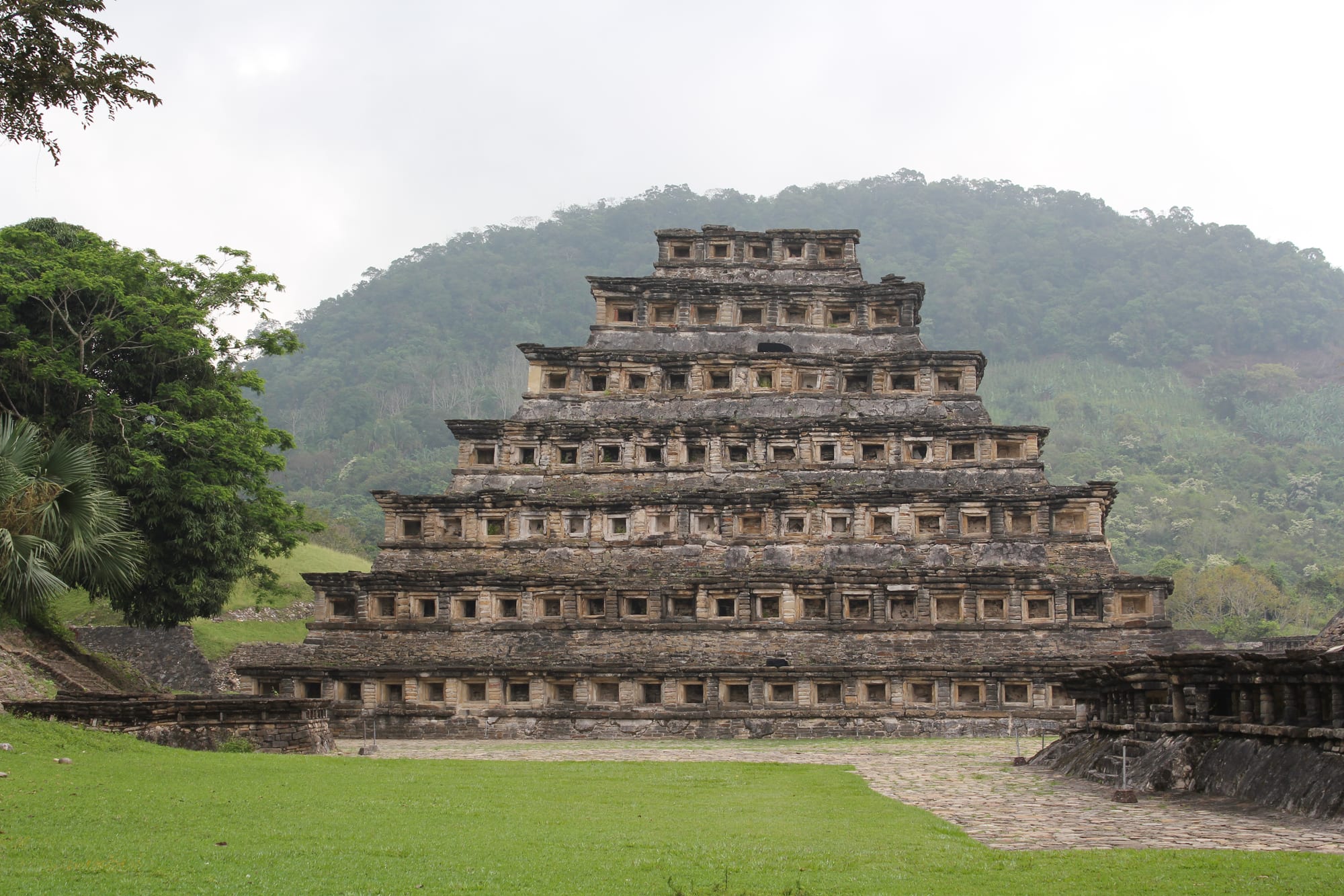 A stepped stone pyramid with rows of square niches on every tier, in front of a forested green hill