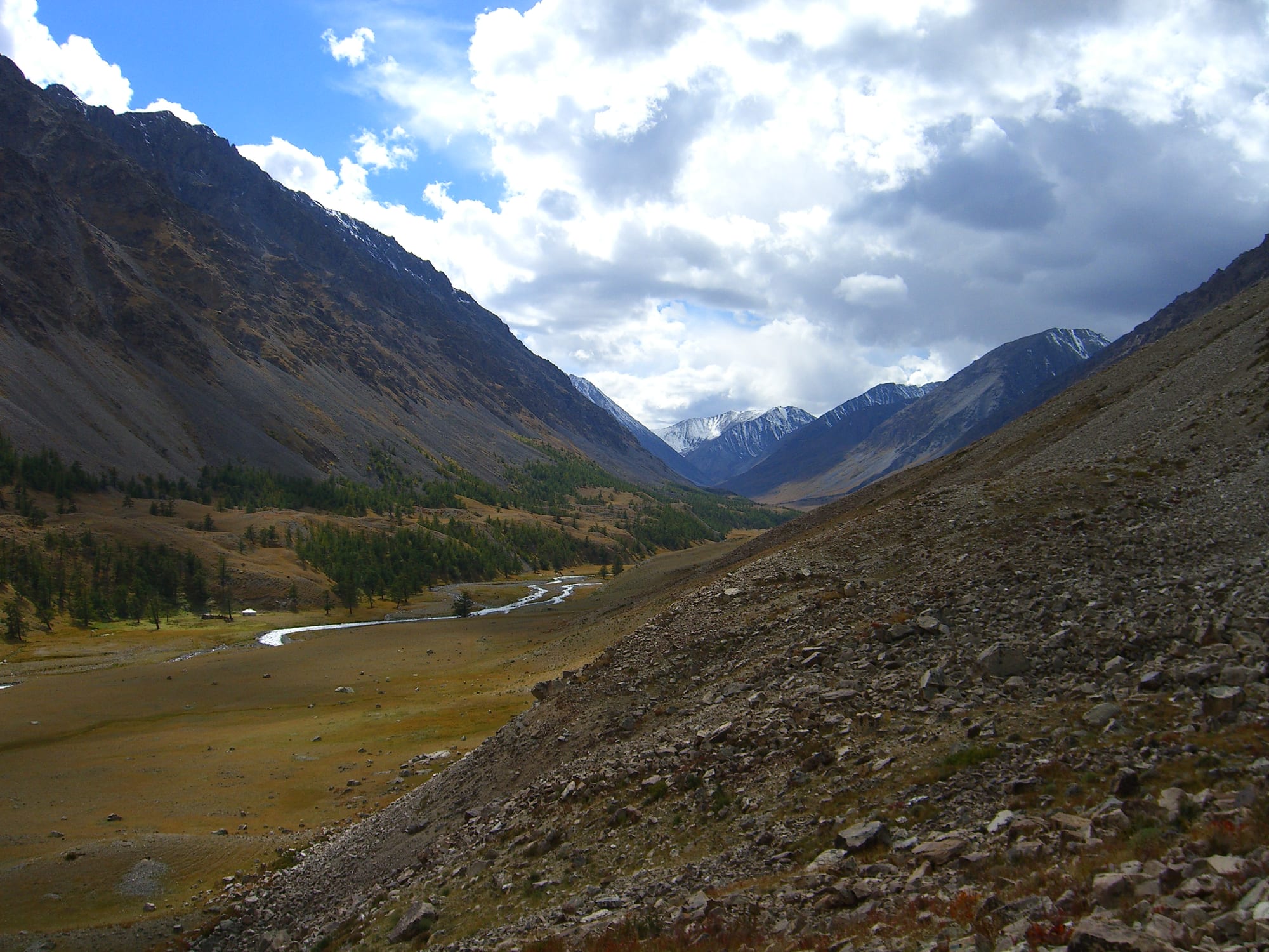 A river winding through a broad valley of larch and autumn grass between steep scree slopes and snowy peaks