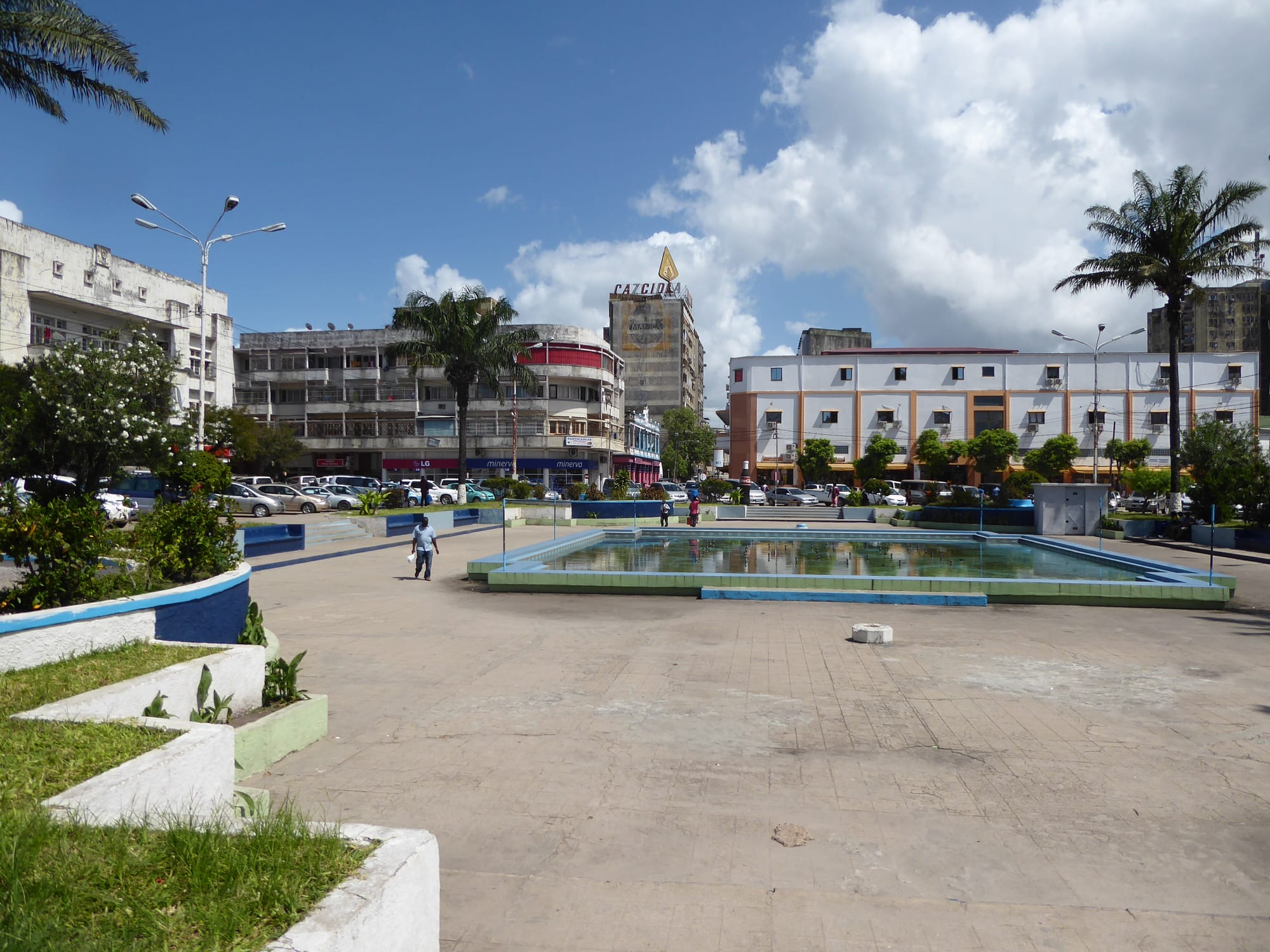 A wide paved square with a shallow rectangular pool, palm trees and flower beds, surrounded by multi-storey concrete buildings under a blue sky