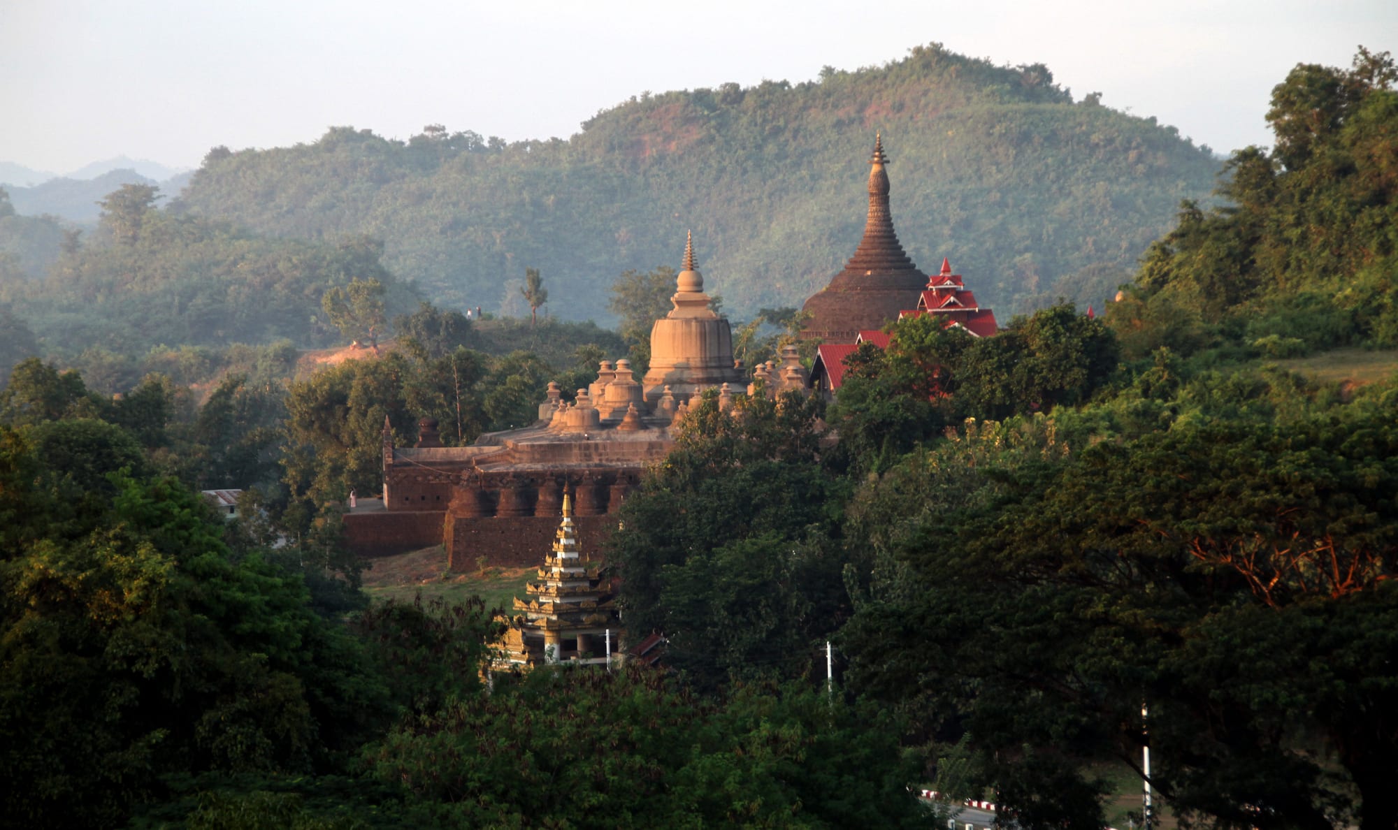 Stone stupas and the walls of an old temple complex among dense green trees, with forested hills behind in soft evening light