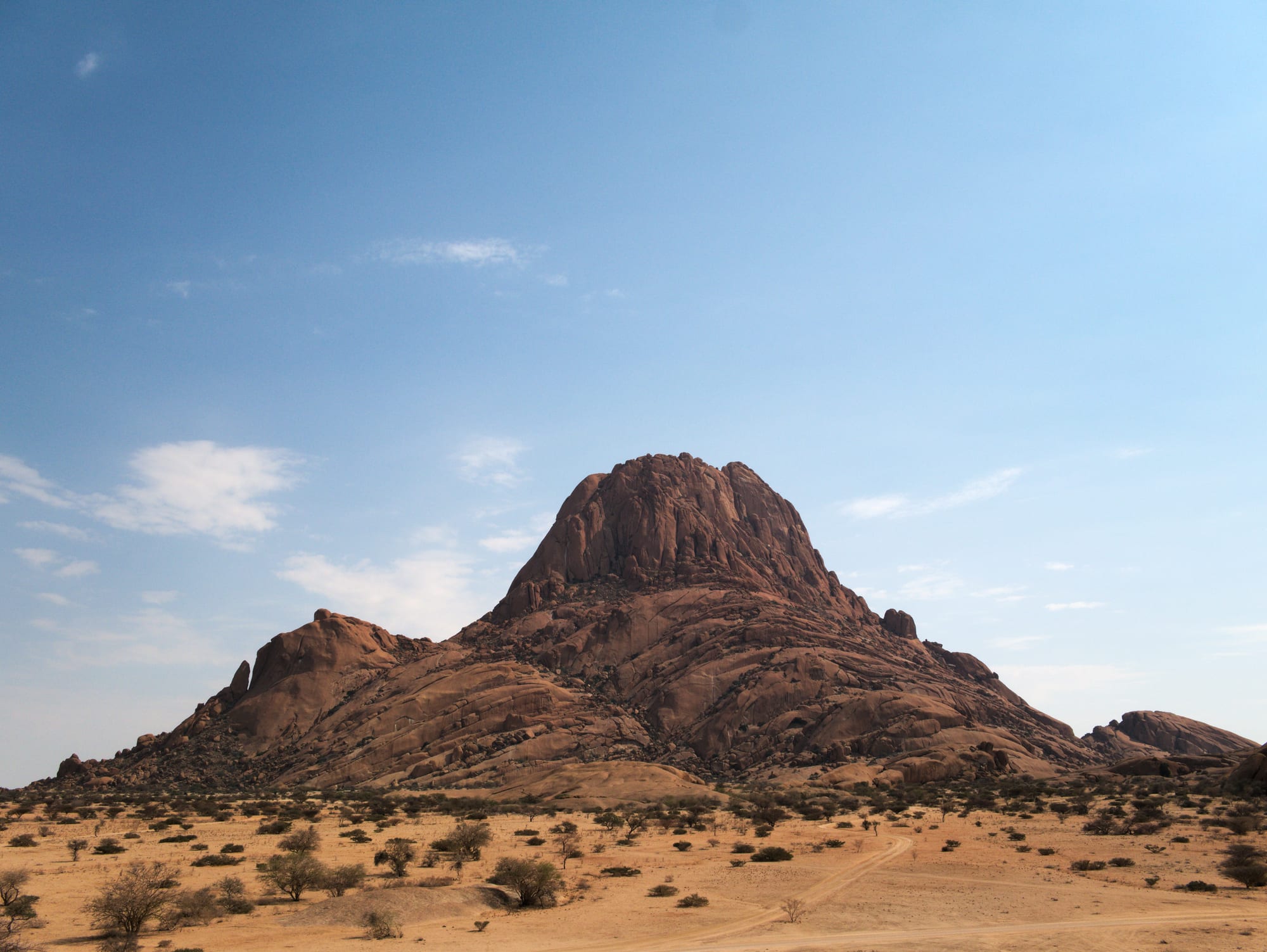 A reddish granite peak rising above flat sandy semi-desert dotted with low shrubs under a blue sky