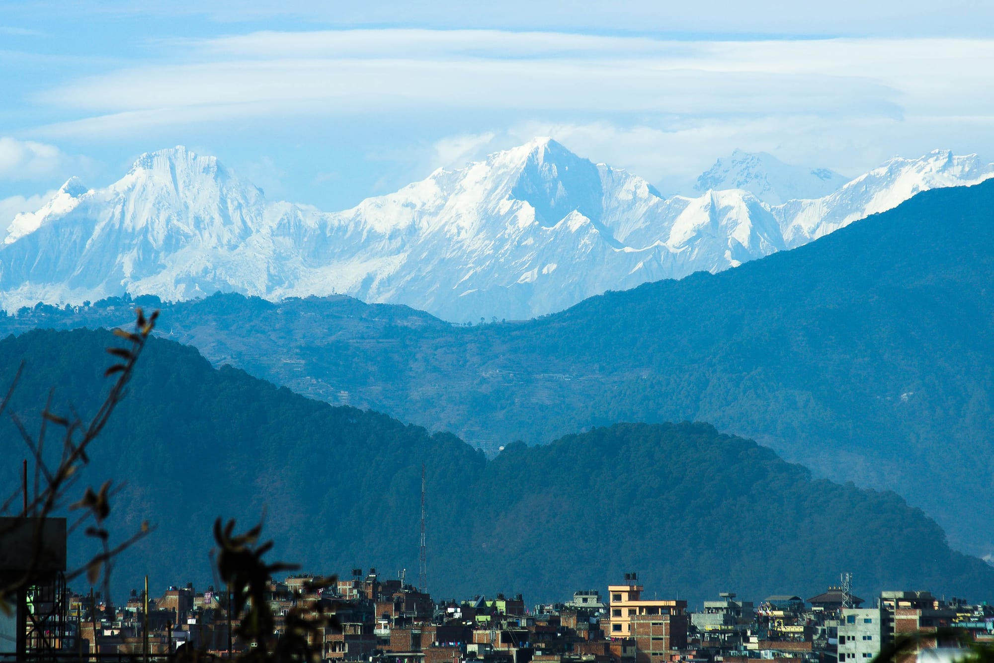 Snow-covered peaks of the Ganesh Himal rising above forested blue ridges, with city rooftops in the foreground