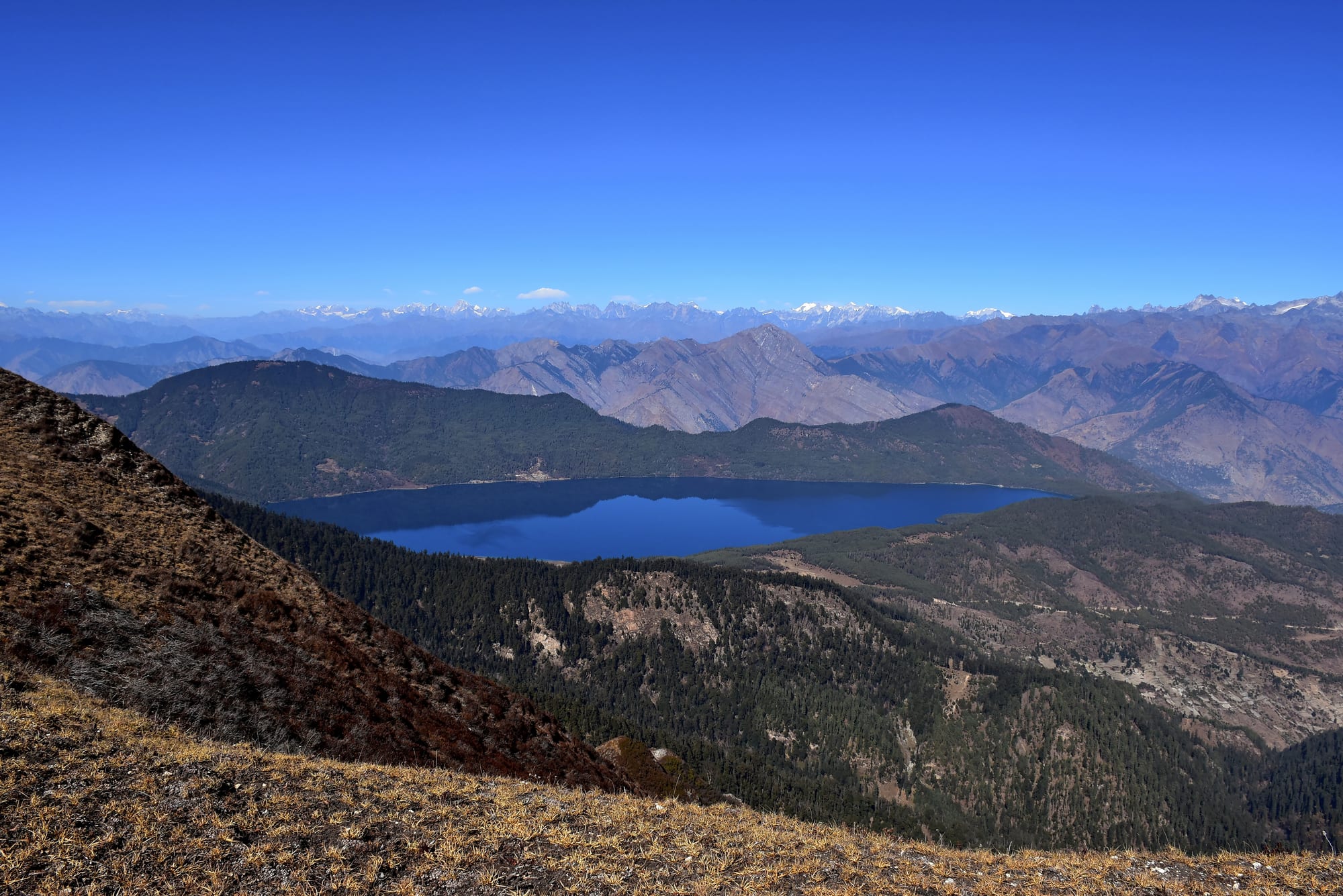 A deep blue lake set among forested ridges, with bare brown hills and a line of snowy peaks beyond under a clear sky