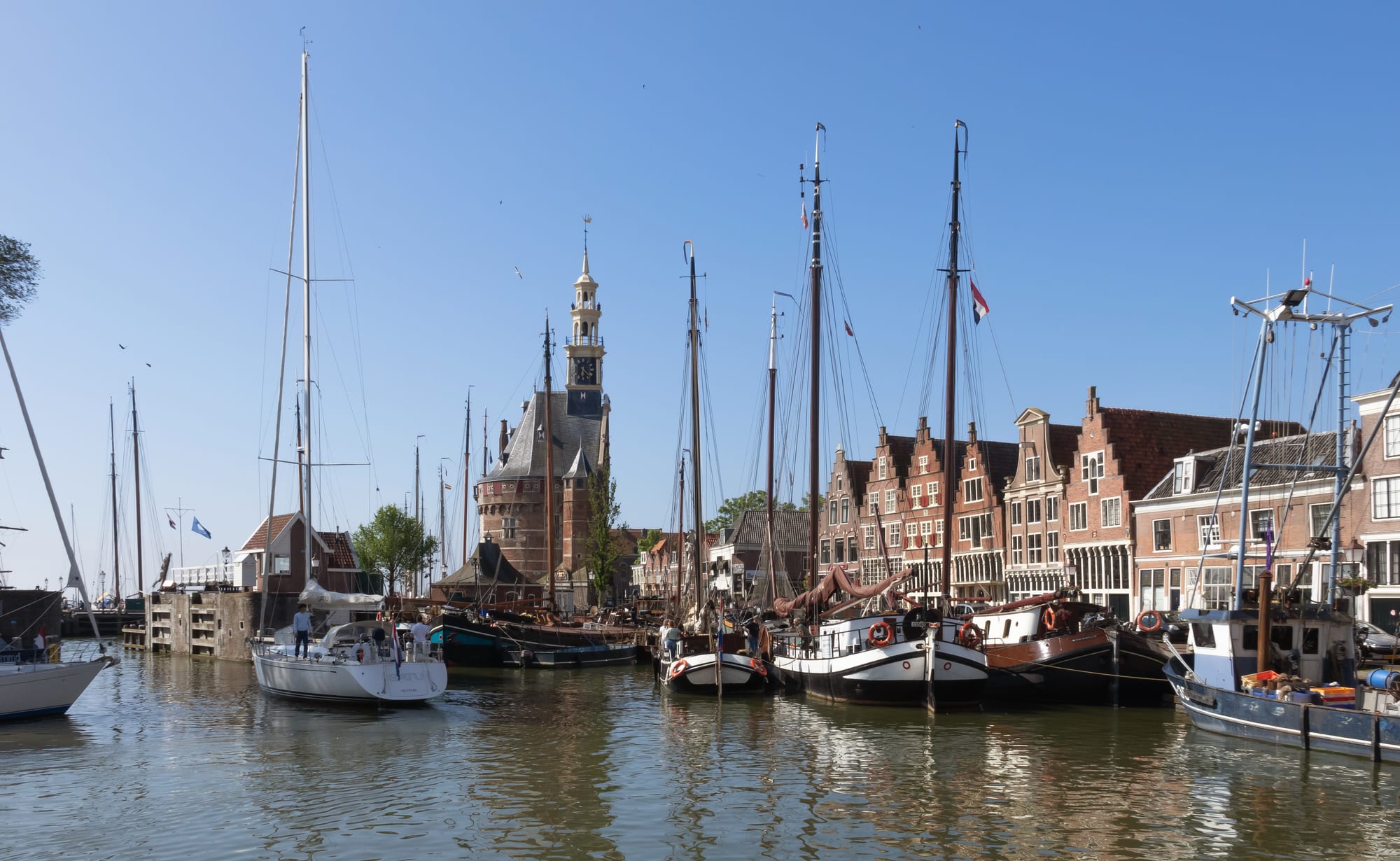Old sailing barges and yachts moored in a calm harbour lined with gabled brick houses, with a brick tower topped by a white wooden spire under a clear blue sky