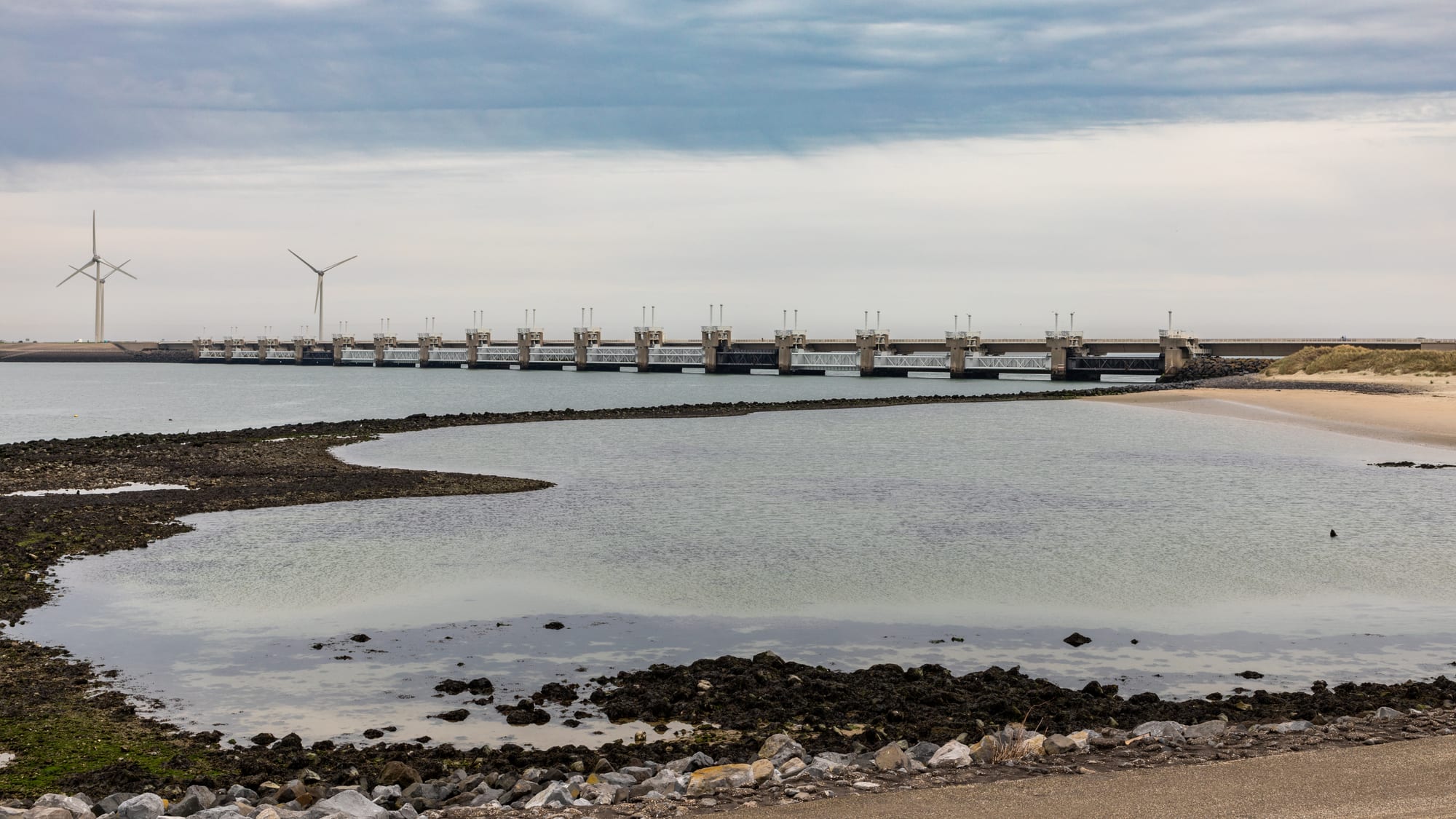 A long storm-surge barrier of concrete piers and steel gates crossing grey water, with sand, rocks and wind turbines