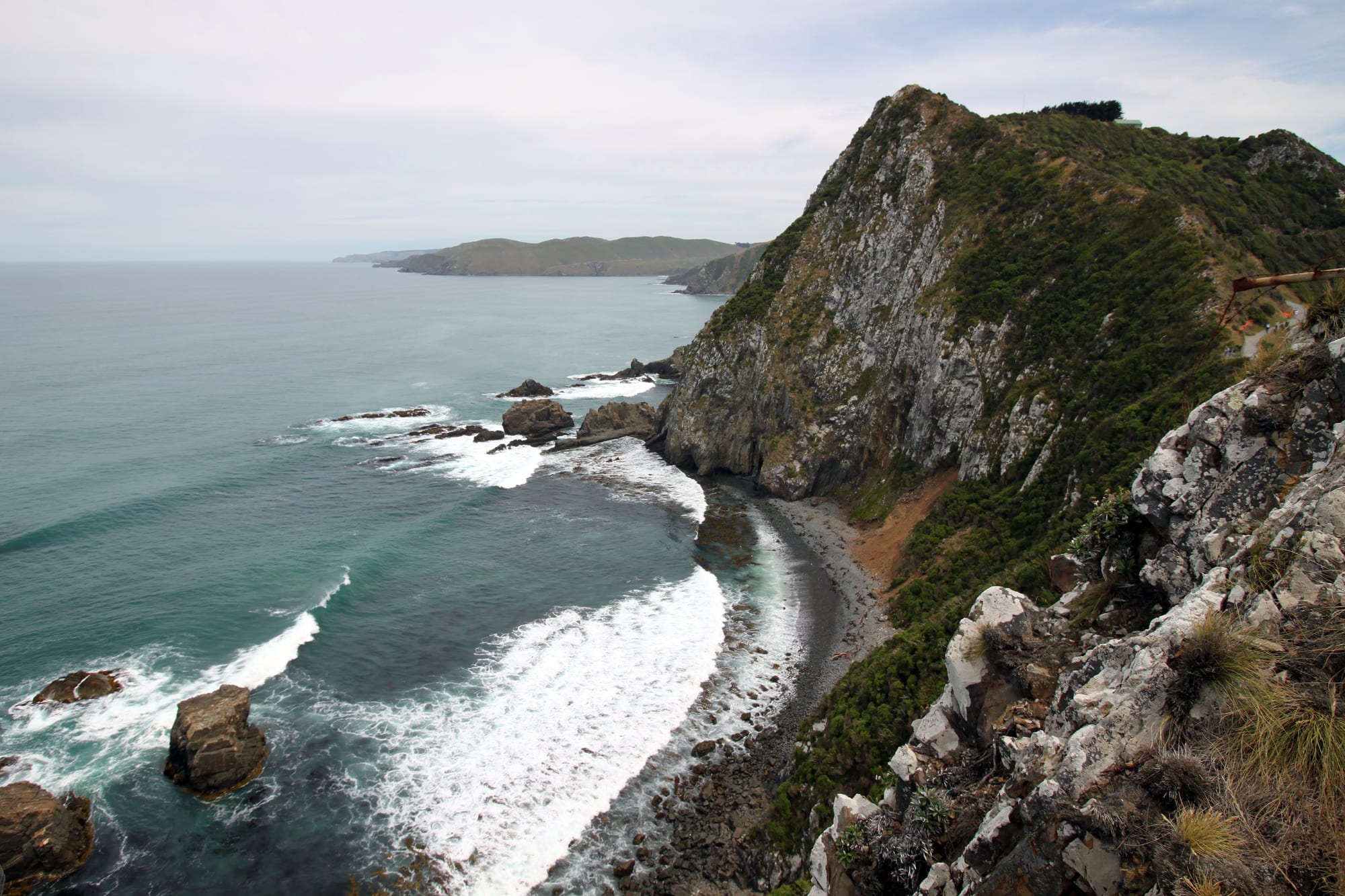 A steep, scrub-covered headland dropping to a curving beach, with rocks and white surf in a green sea