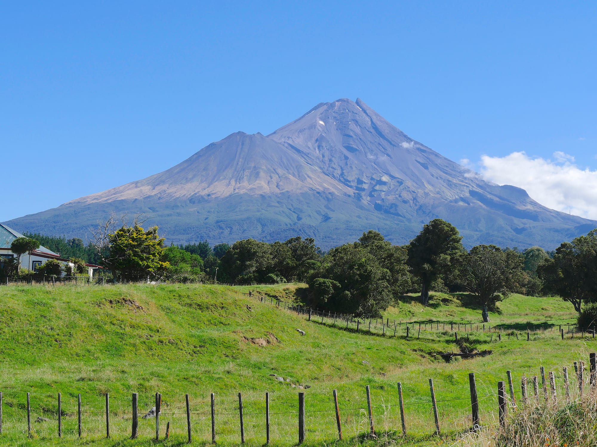 A large cone-shaped volcano under a clear blue sky, rising above green fenced farmland and trees
