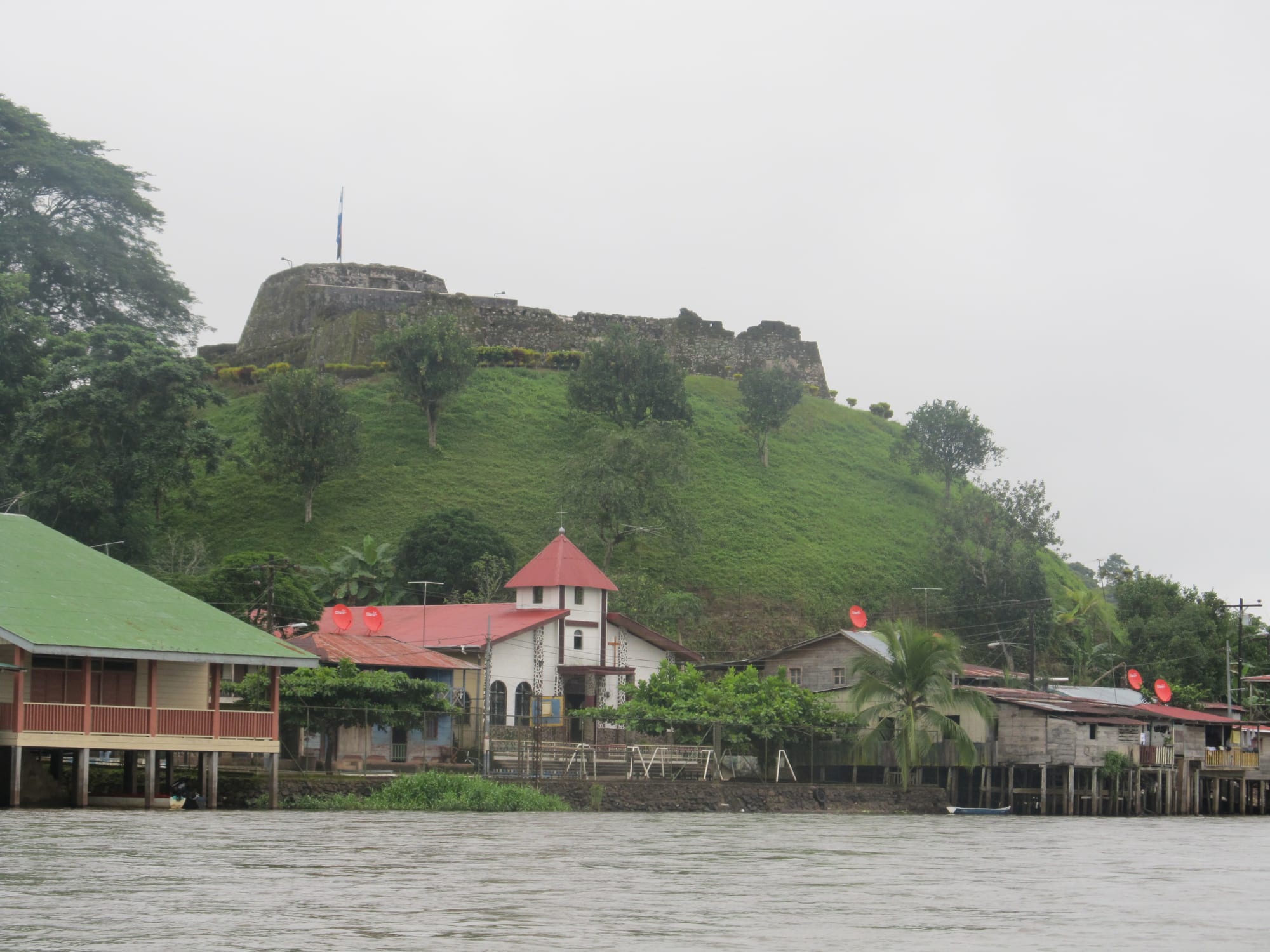 A stone fortress on a green hill above a riverside village with a church and stilt houses along the water