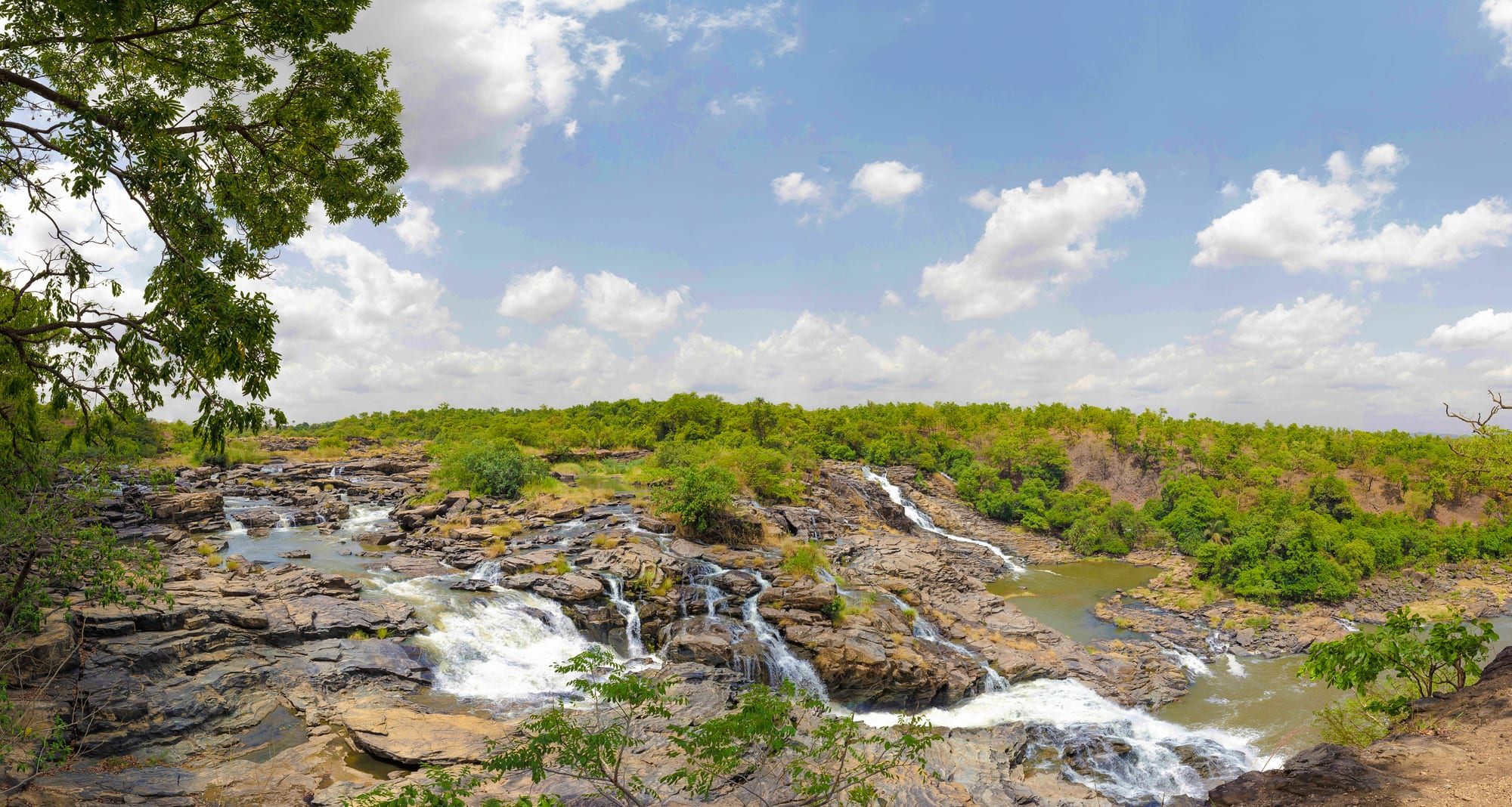 Water cascading over wide shelves of dark rock into pools, surrounded by green bush under a blue sky with white clouds