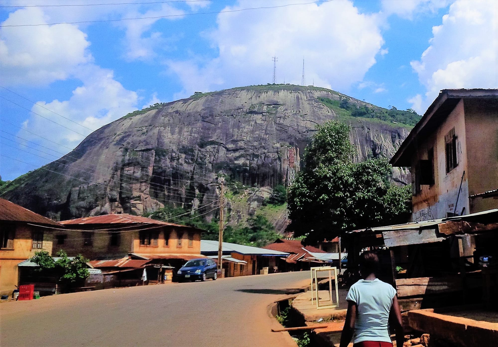 A huge bare granite dome rising behind houses with rusty roofs and a curving road under a blue sky with white clouds