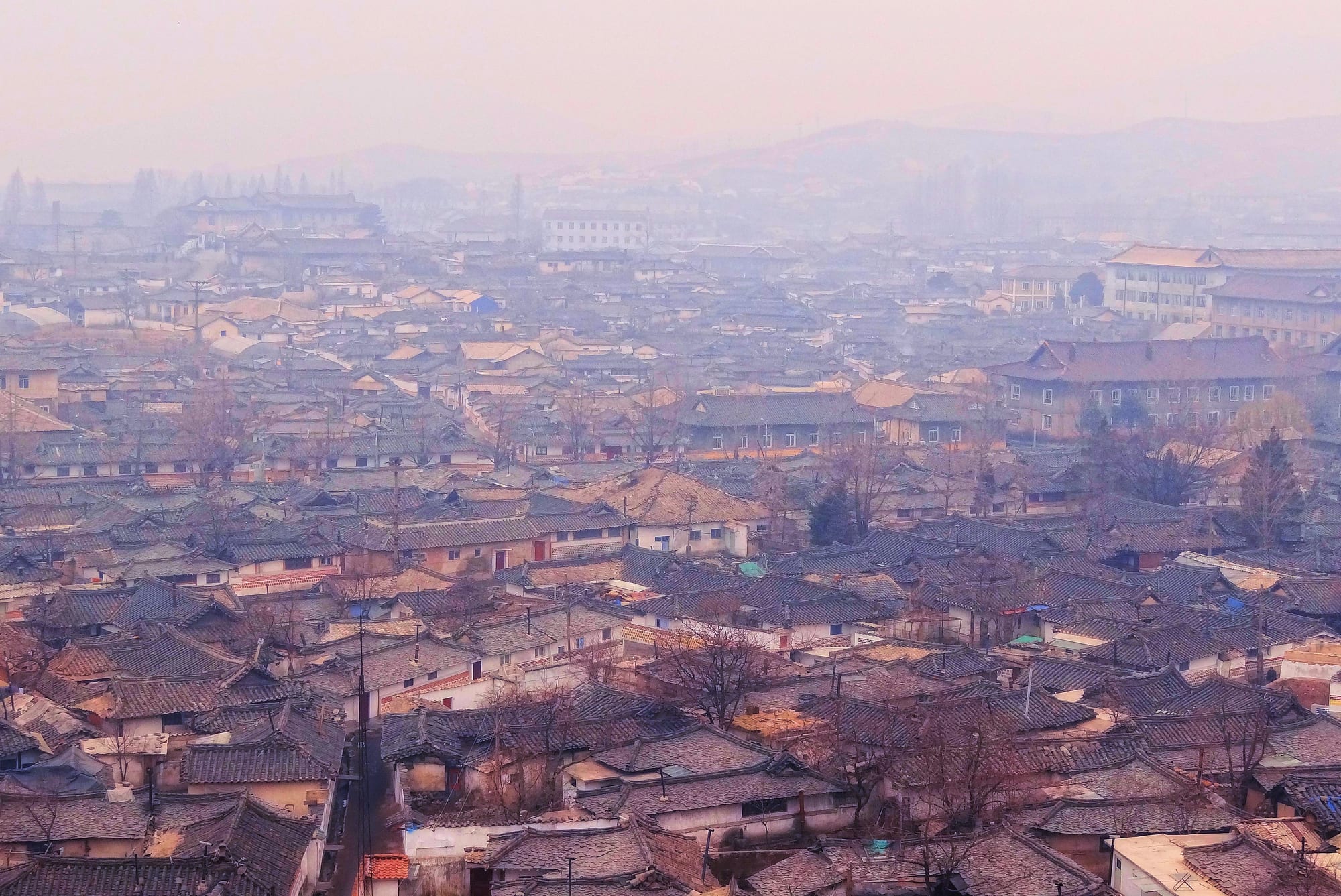 Hazy view over a dense spread of grey tiled traditional roofs with larger buildings and hills in the distance