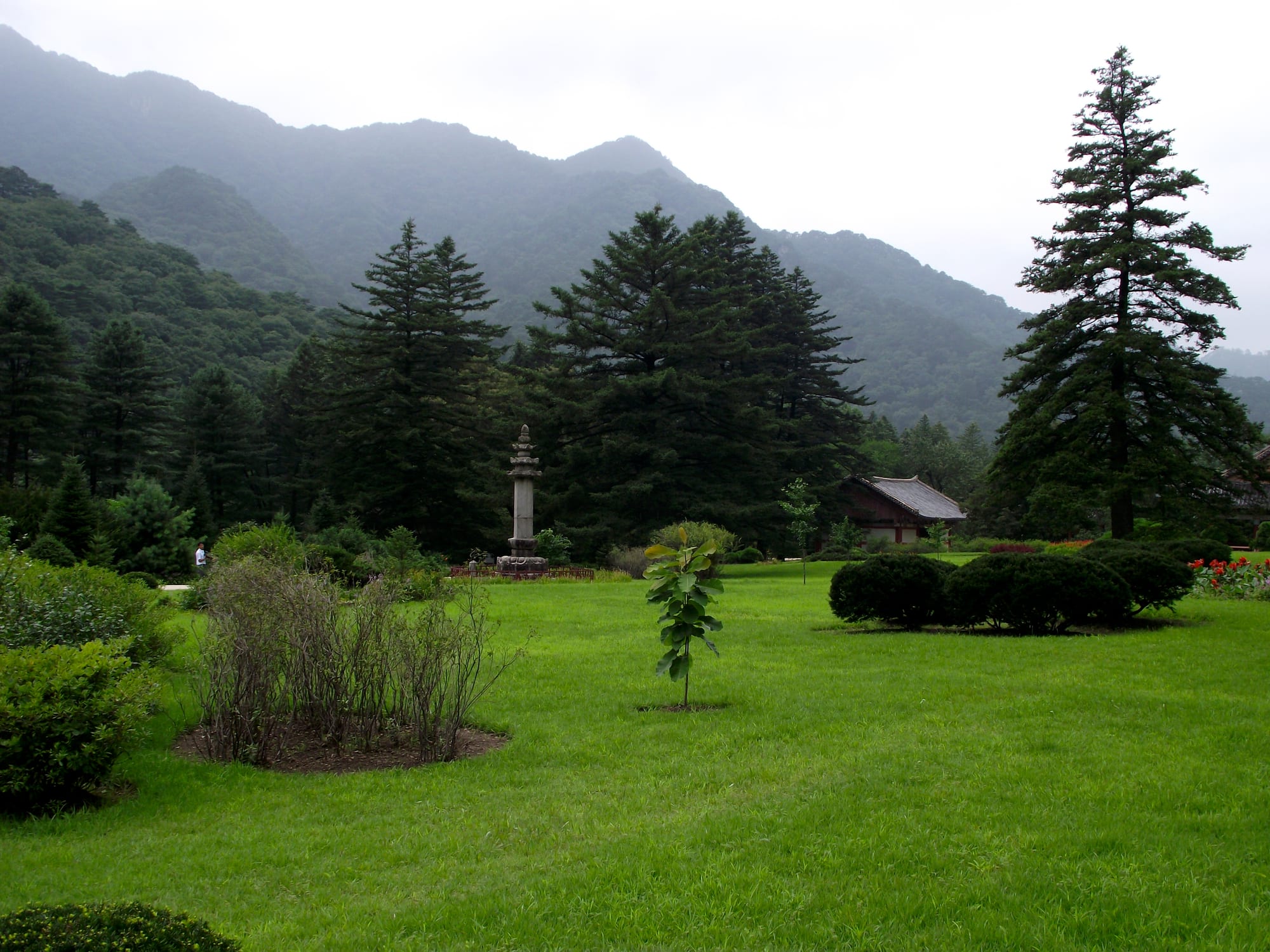 A green lawn with a stone pagoda and tall conifers in front of forested, mist-covered mountain ridges