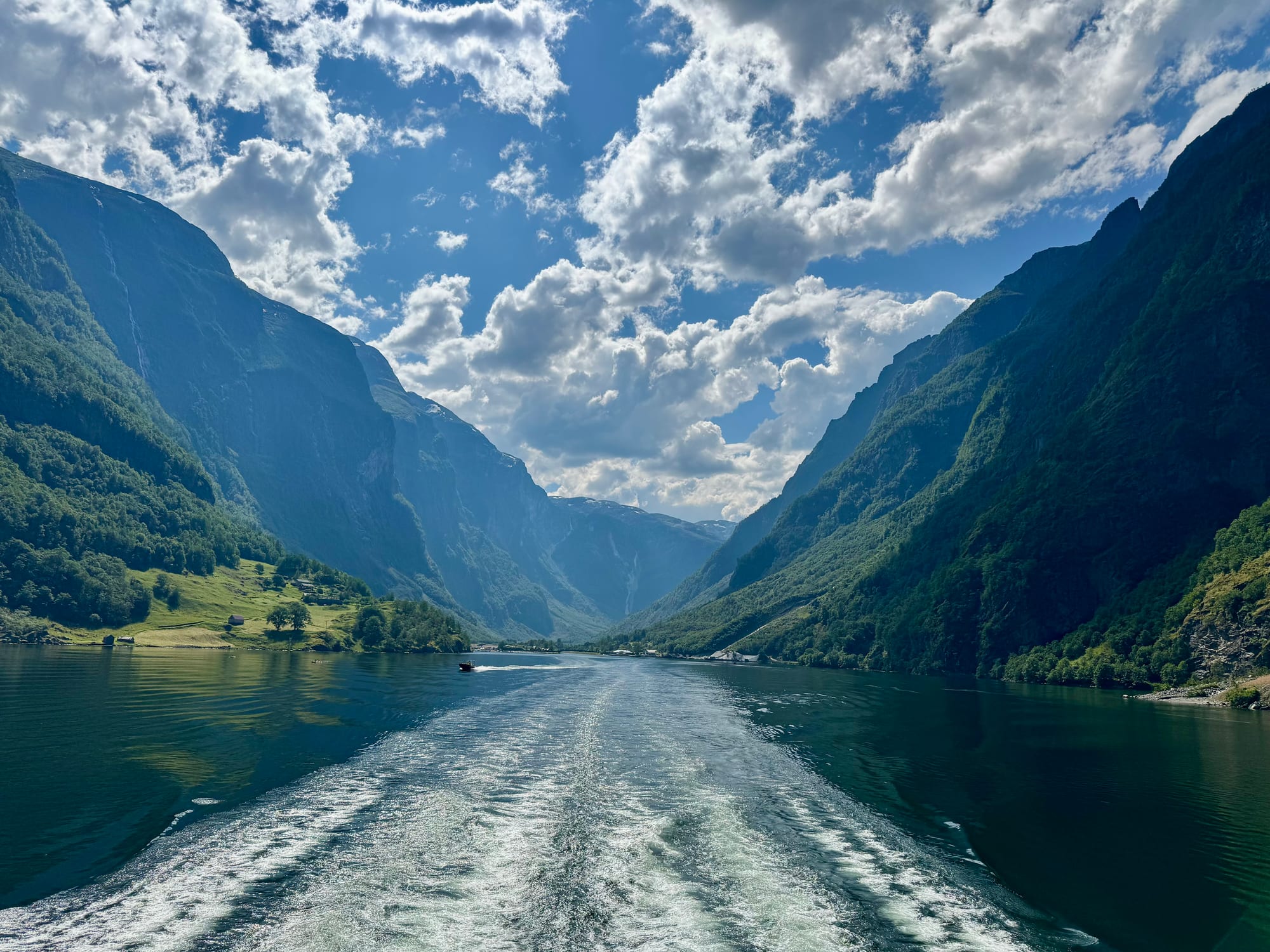 A narrow fjord between steep green mountainsides under a cloudy blue sky, with a boat's white wake in the foreground