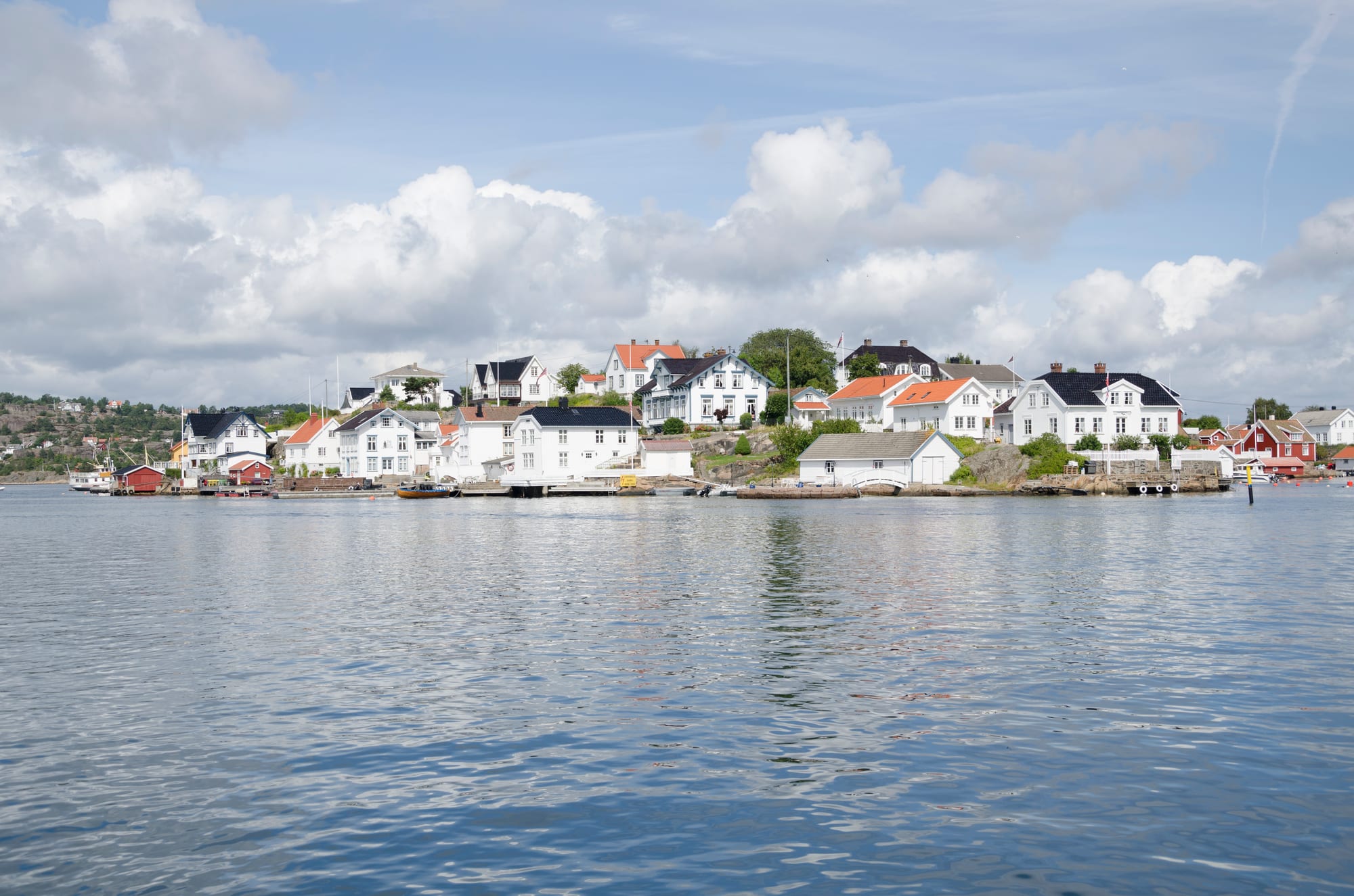 White-painted wooden houses and boathouses along the shore of a small island, seen across calm water under summer clouds