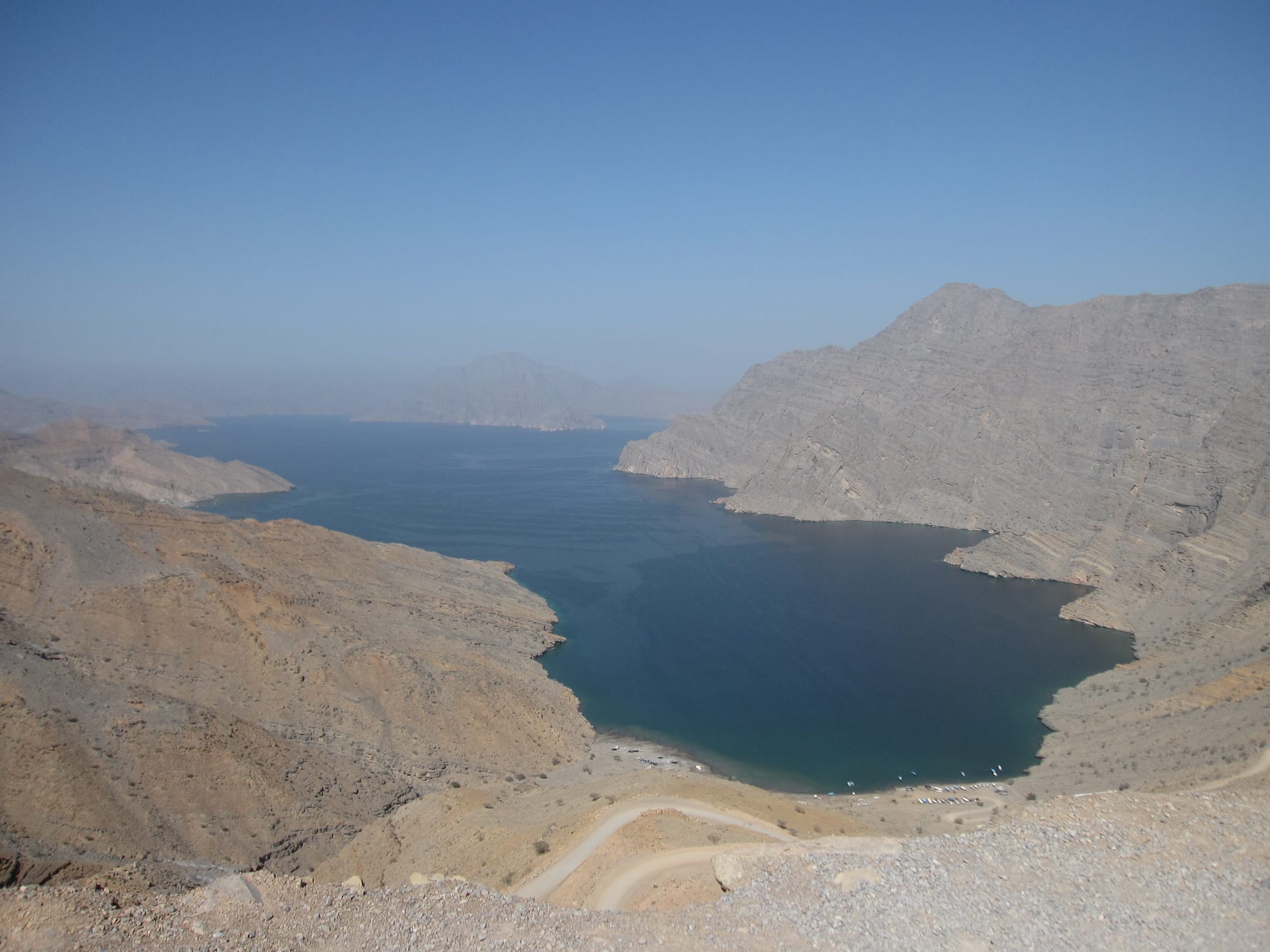 A deep blue inlet between steep, bare grey mountain slopes, seen from a hillside road above a small beach