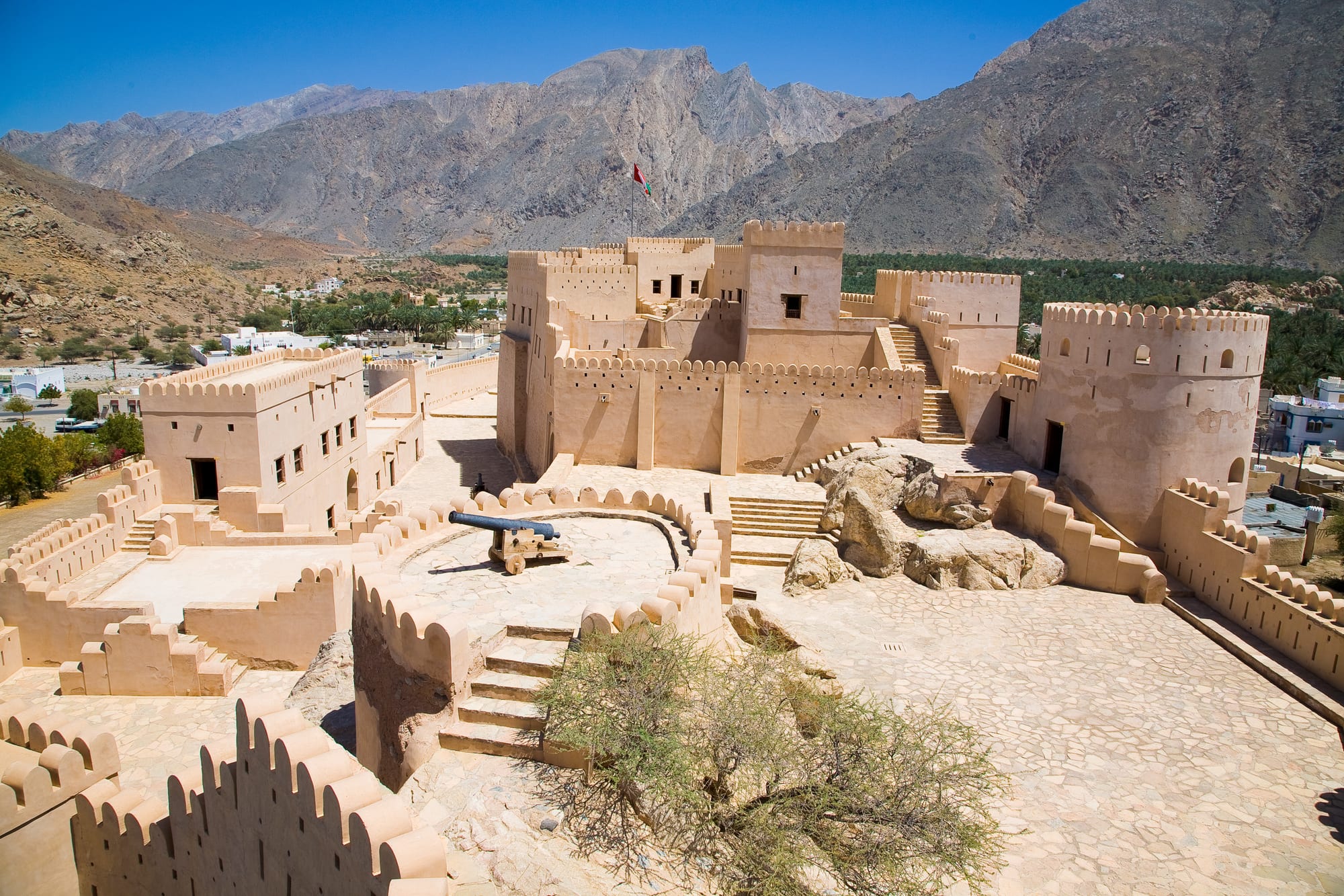 Sand-coloured crenellated walls and round towers of Nakhal Fort, with a palm oasis and bare grey mountains behind